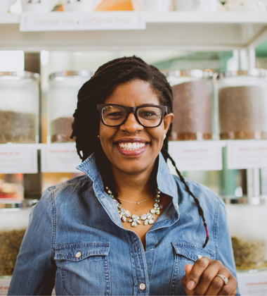 Woman wearing glasses and a denim shirt smiling in front of shelves with jars of herbs and spices.