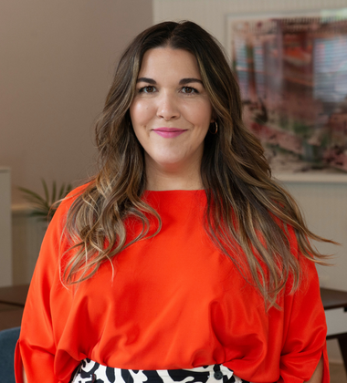 A woman with long wavy hair wearing a bright orange top and patterned skirt, smiling indoors.