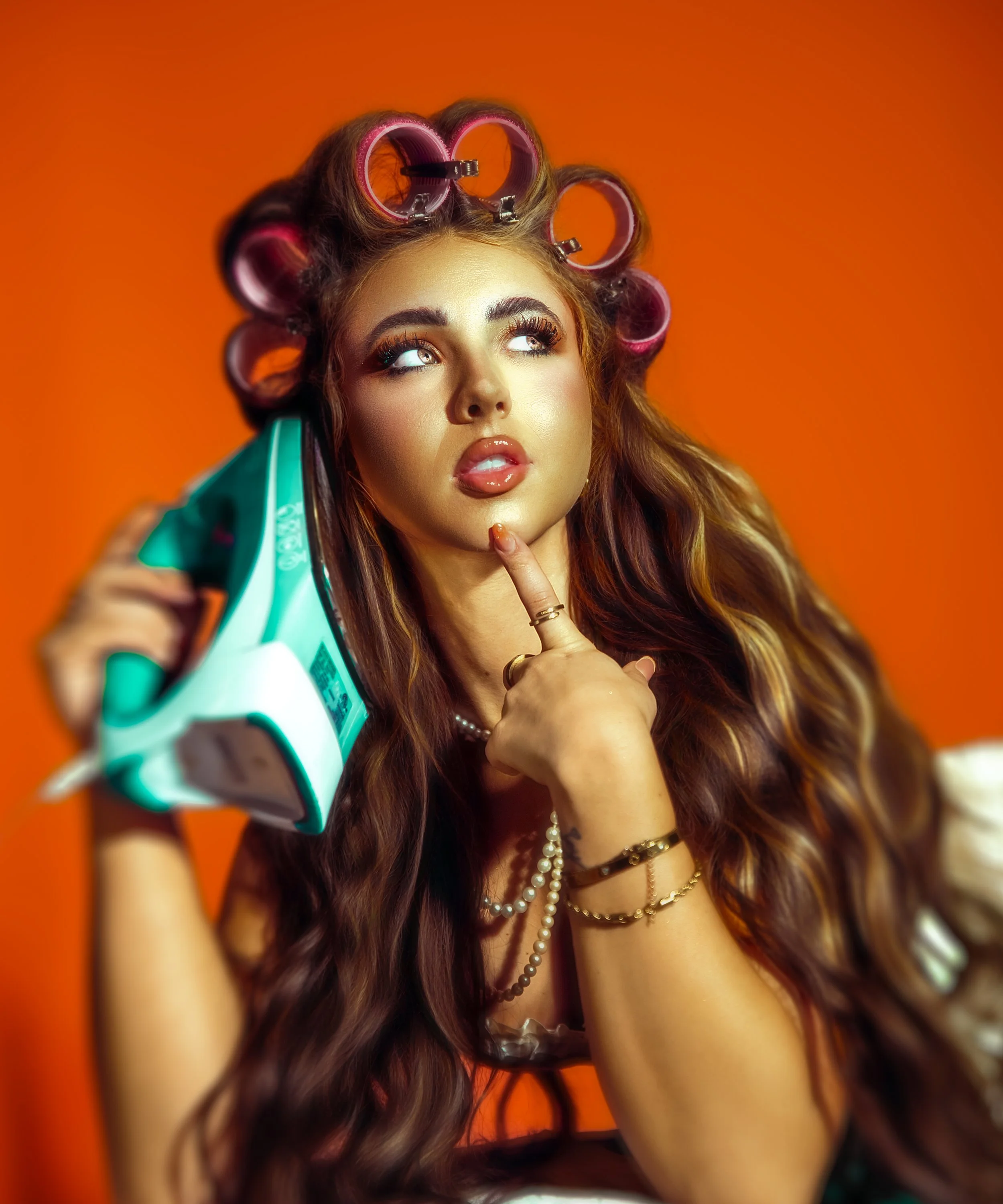 A woman with long wavy brown hair is holding a box of hair rollers in front of her face, with some rollers in her hair. Long eyelashes and glossy lips, wearing jewelry pearls and bracelets. The background is orange. Cleveland Photographer. Cleveland 