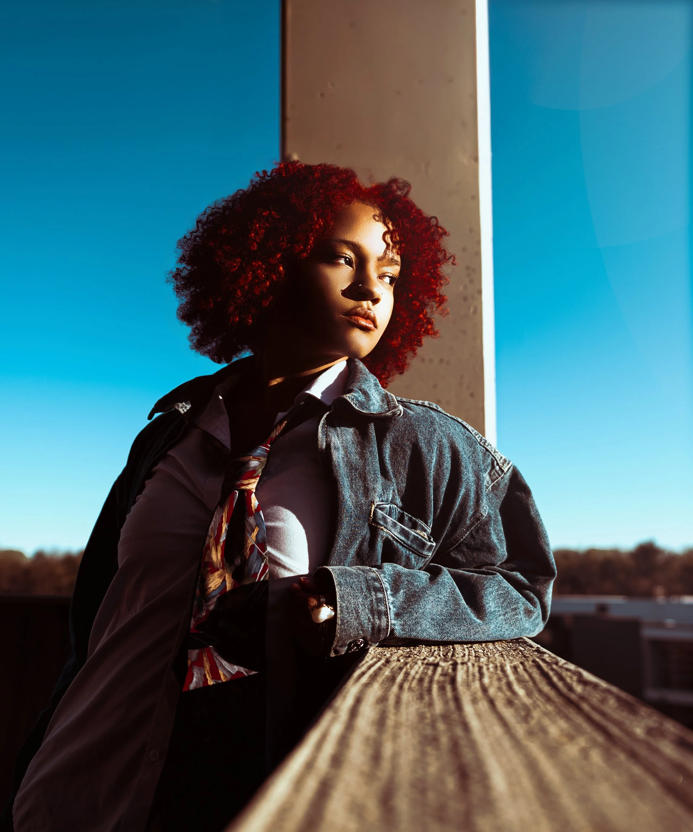 A young woman with curly red hair standing outdoors by a wooden railing, with blue sky and horizon in the background, looking to her left. Cleveland Photographer. Cleveland Portrait Photographer, Legacy Village