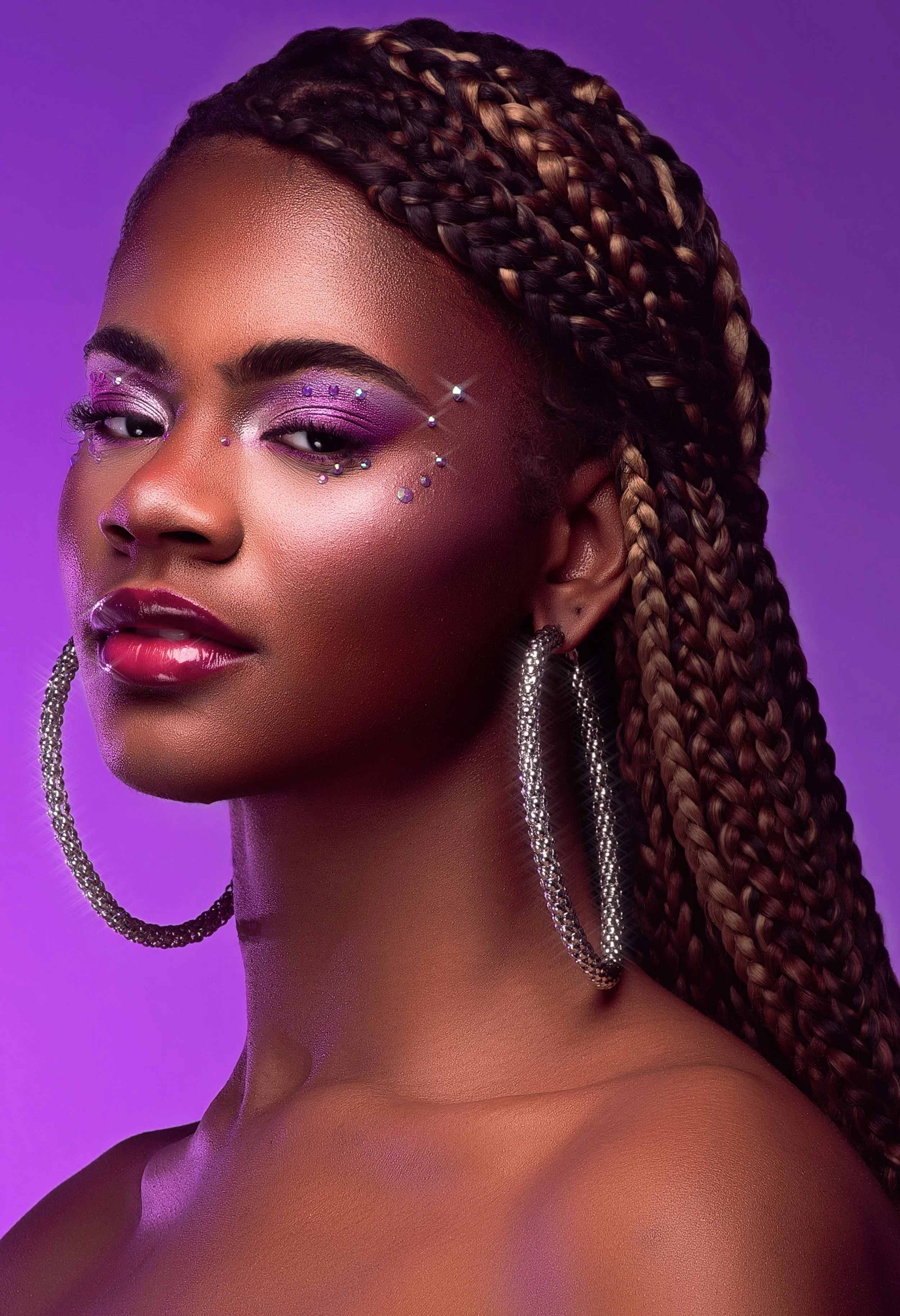 A close-up portrait of a woman with braided hair, vibrant purple eyeshadow, rhinestones around her eyes, large hoop earrings, and a purple background. Cleveland Photographer. Cleveland Portrait Photographer
