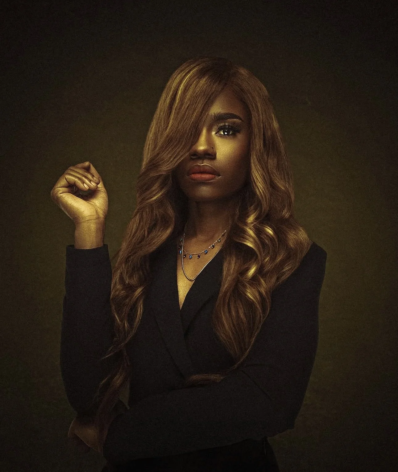 A woman with long, wavy light brown hair and dark skin wearing a black blazer, gold makeup, and colorful jewelry, posing against a dark background. Cleveland Photographer. Cleveland Portrait Photographer