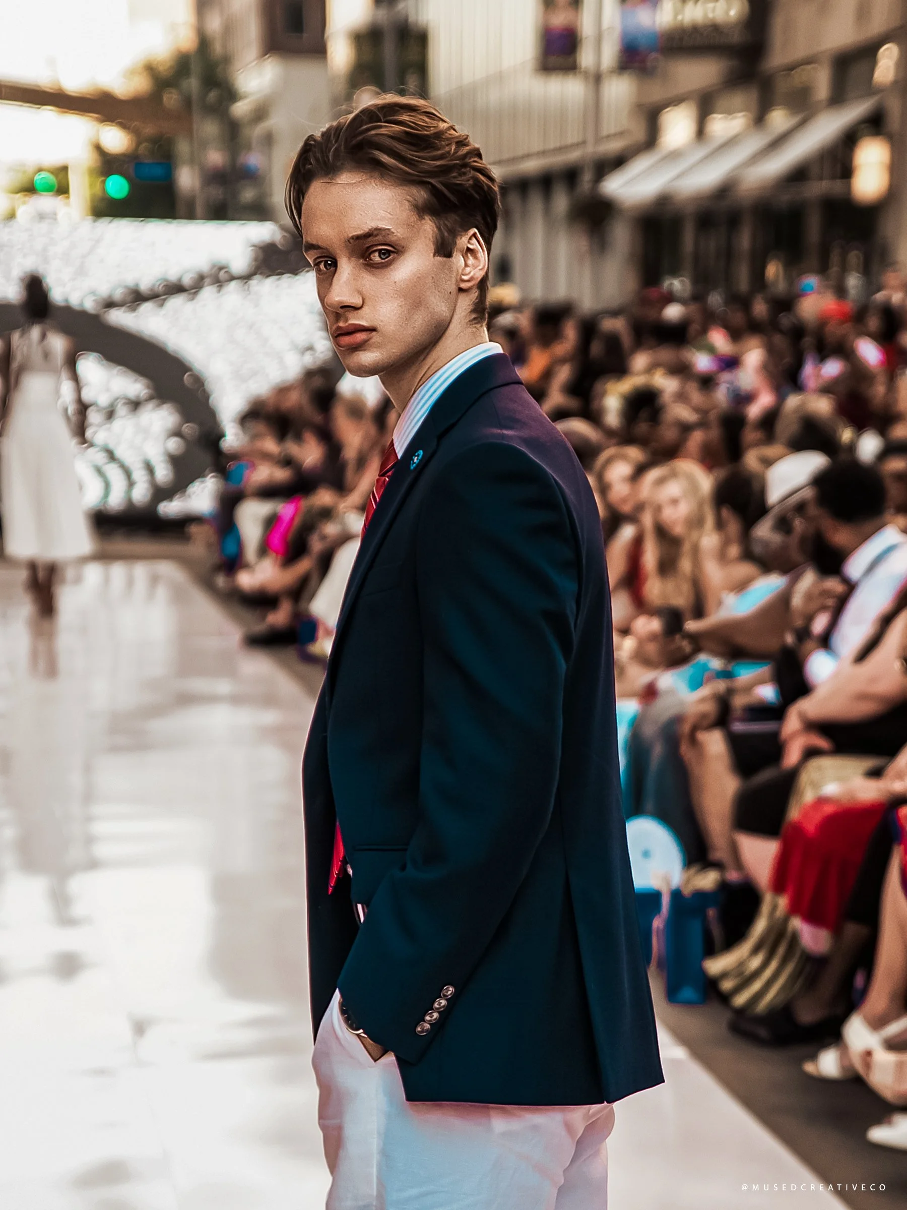 Young man with a dillards navy blazer with metallic buttons on the sleeve, white shirt with blue stripes, red tie, and white pants, looking over his shoulder in a busy retail store or mall. Cleveland Photographer. Cleveland Portrait. Fashion Talks