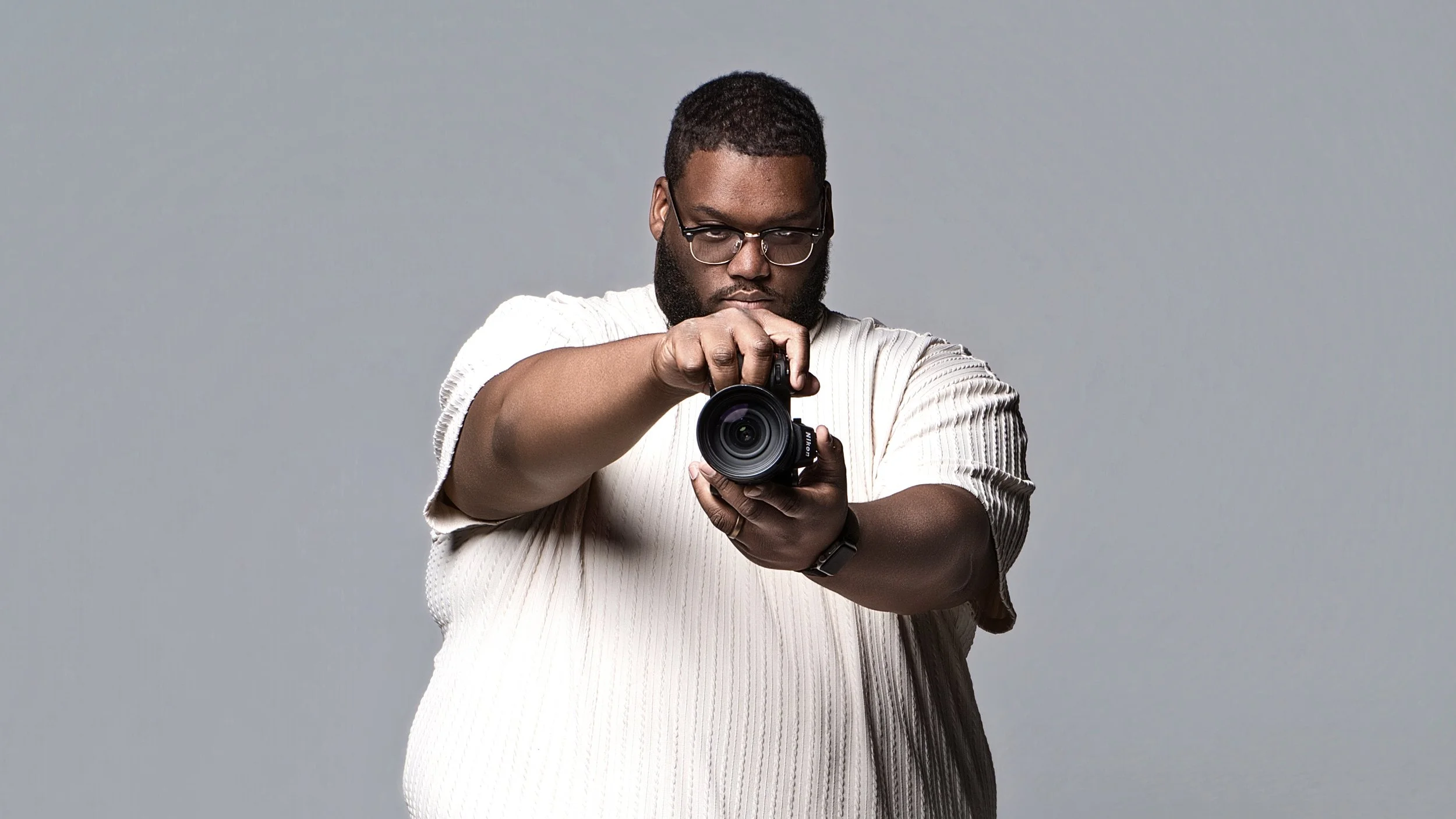 Derick Jerome or Derick Todd with glasses and a beard holding a camera, pointing it towards the viewer, against a plain gray background. Cleveland Photographer. Cleveland Portrait Photographer