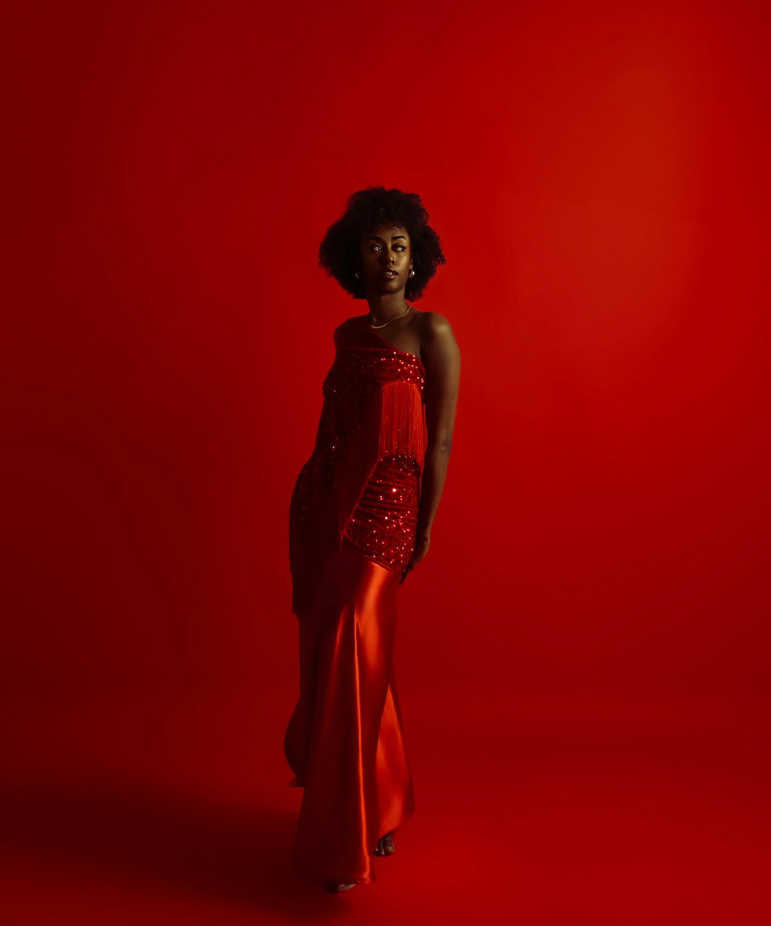 A woman with dark curly hair and dark skin is standing in front of a red background. She is wearing a strapless, red, sequined dress and a gold necklace. She has earrings and is looking to her left. Cleveland Photographer. Cleveland Portrait 