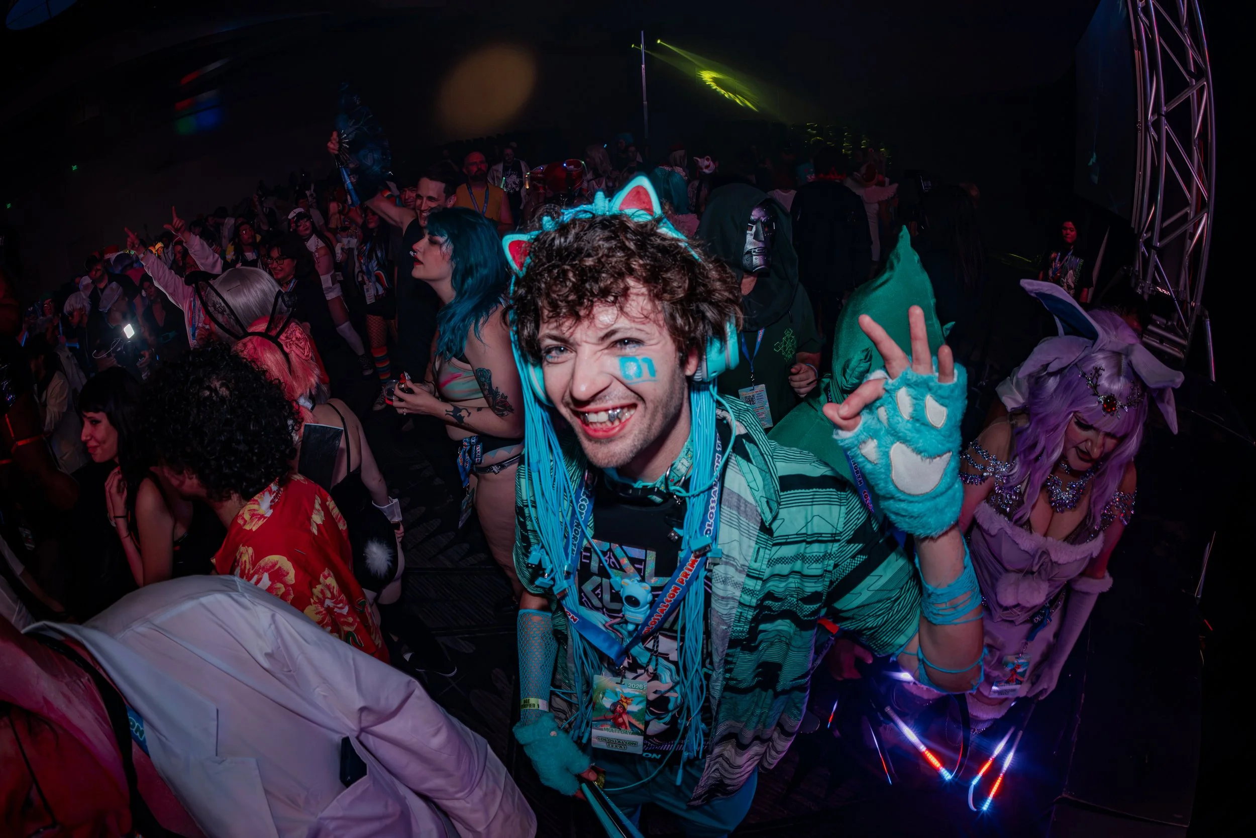 People in colorful costumes and accessories at a lively party or celebration, with dark lighting and laser effects.