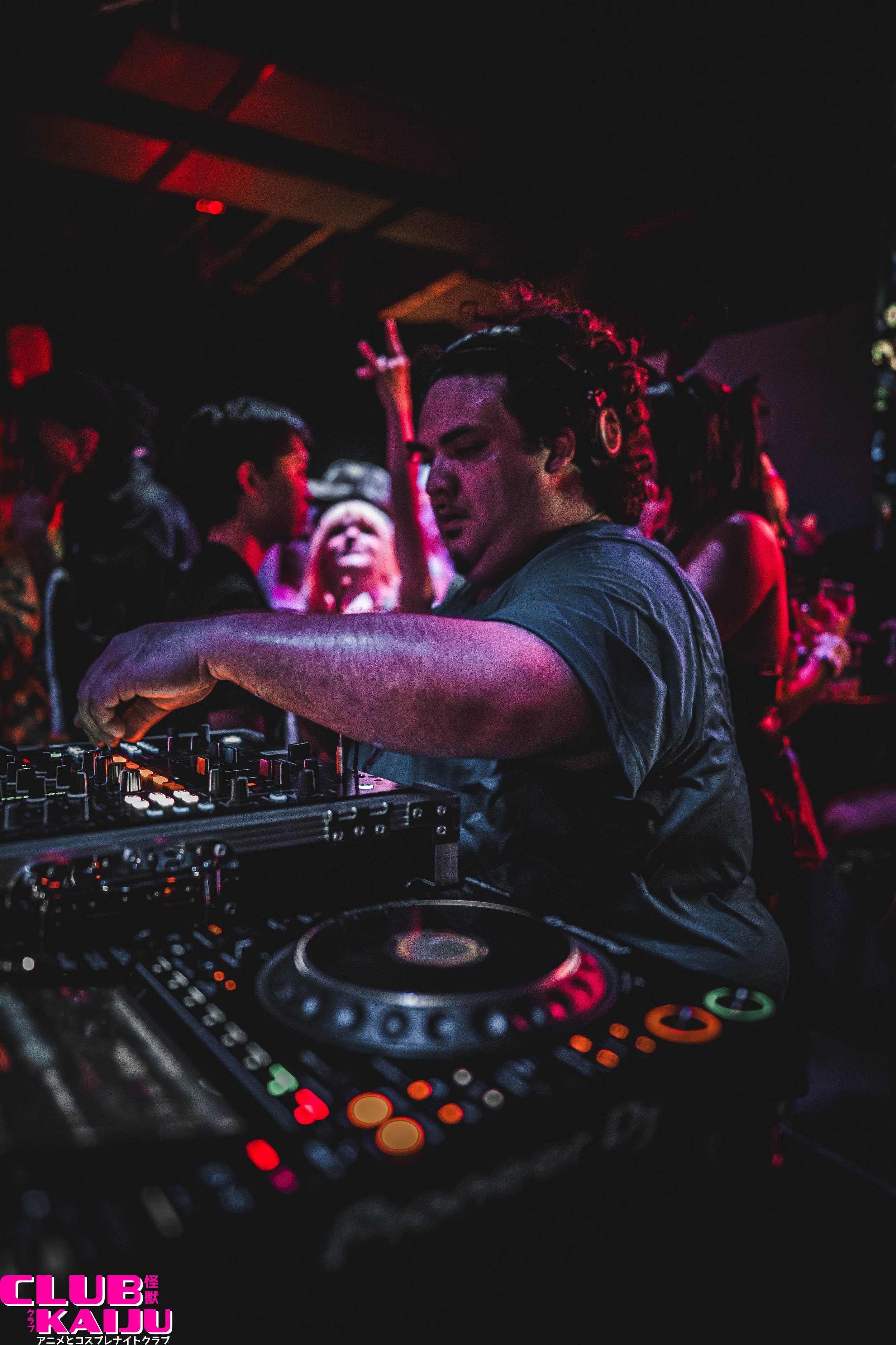 A DJ performing at a nightclub, with a crowd dancing in the background, illuminated by colorful lights, and the logo of Club Kaiji in pink at the bottom left corner.