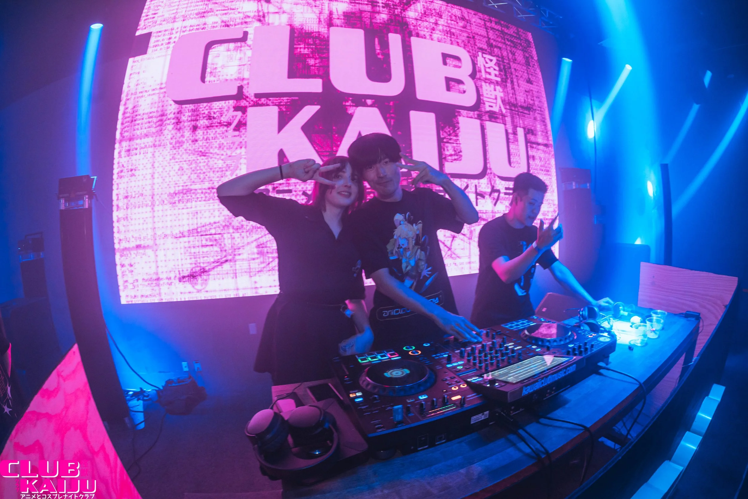 People DJing at a nightclub with a large pink screen reading 'CLUB KAU' in the background, illuminated by blue and purple lights.