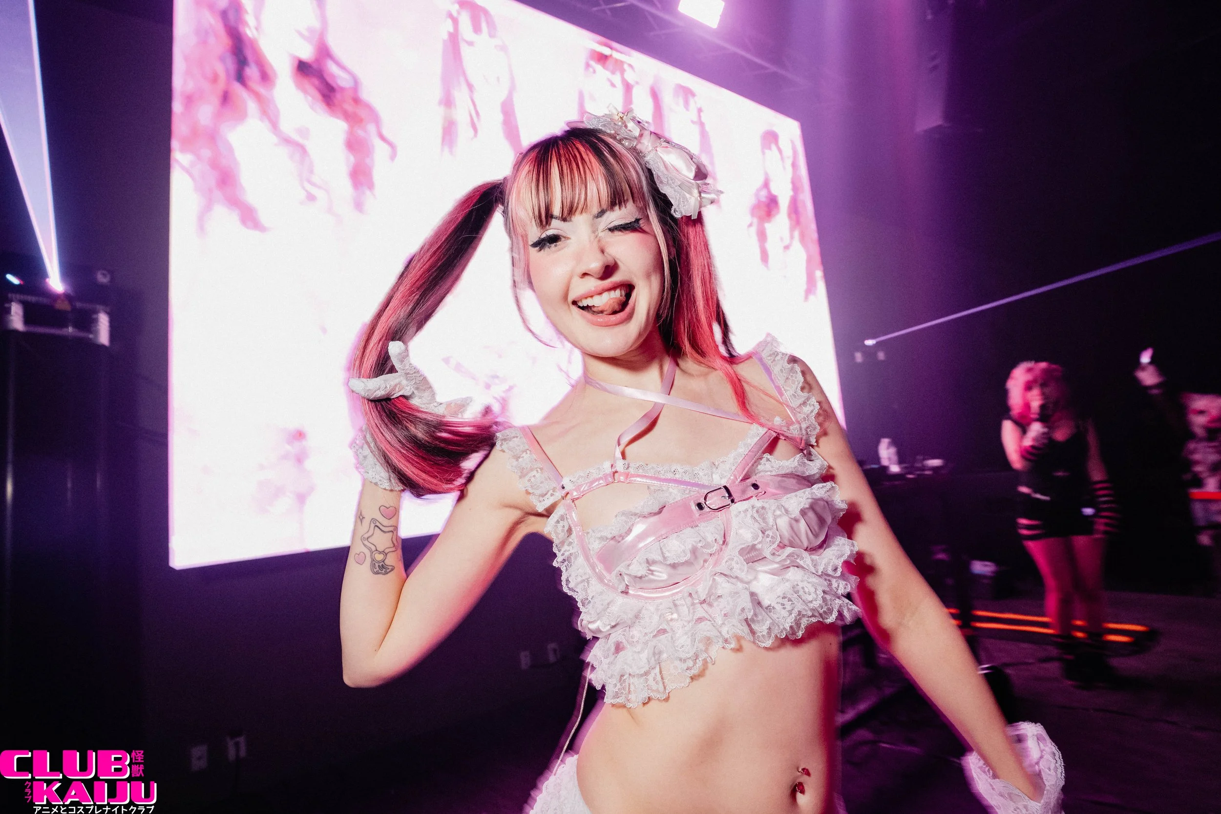 A young woman with pink hair styled in pigtails, wearing a revealing pink and white lace outfit with bows, posing and sticking out her tongue at a club or event. There is a large screen in the background with animated characters and the text "CLUB KAIJU" in the bottom left corner.