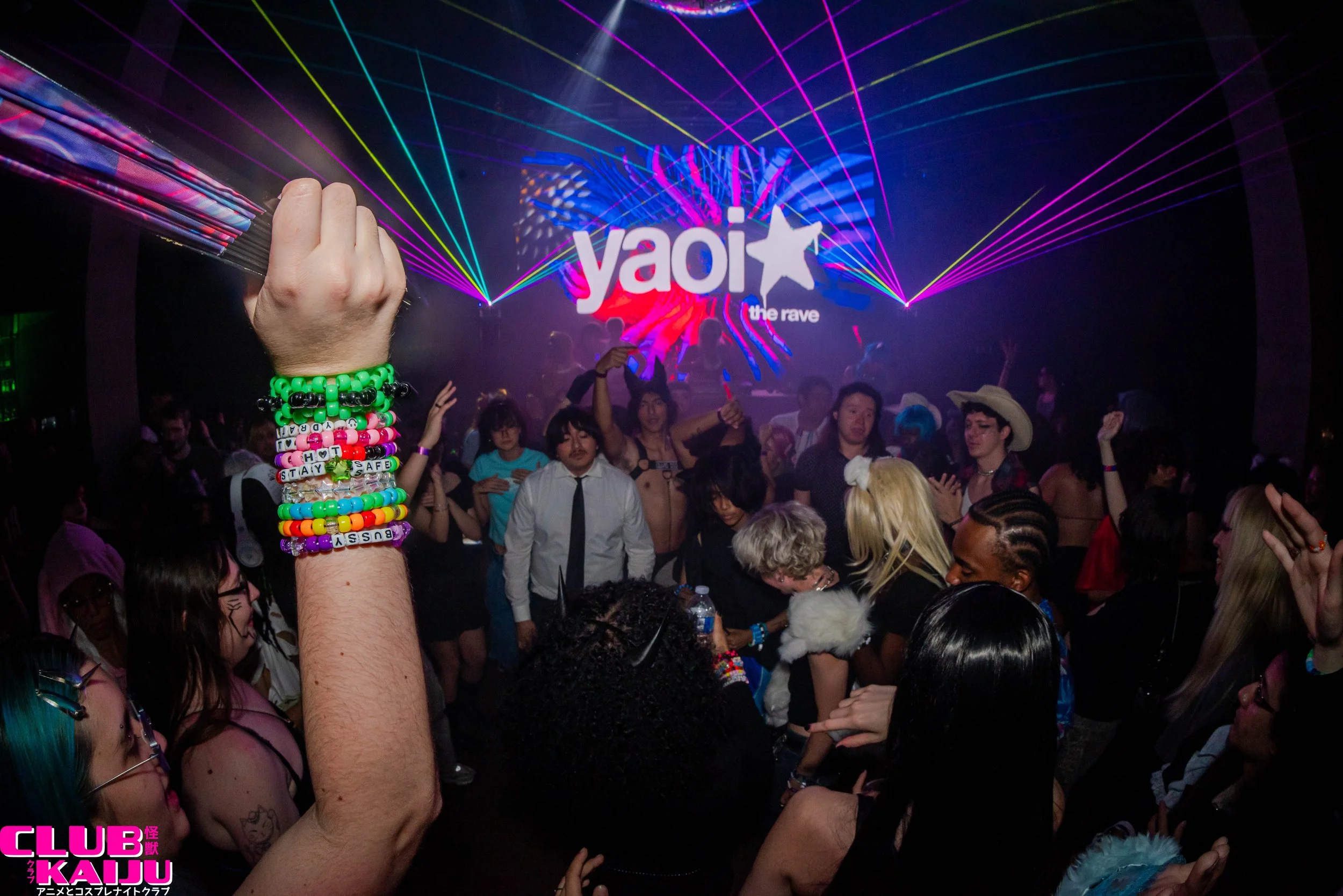 A crowded dance club with colorful laser lights and a large screen displaying the logo 'YO!Y' and the words 'the rave'. An arm with multiple brightly colored beaded bracelets is raised in the foreground.