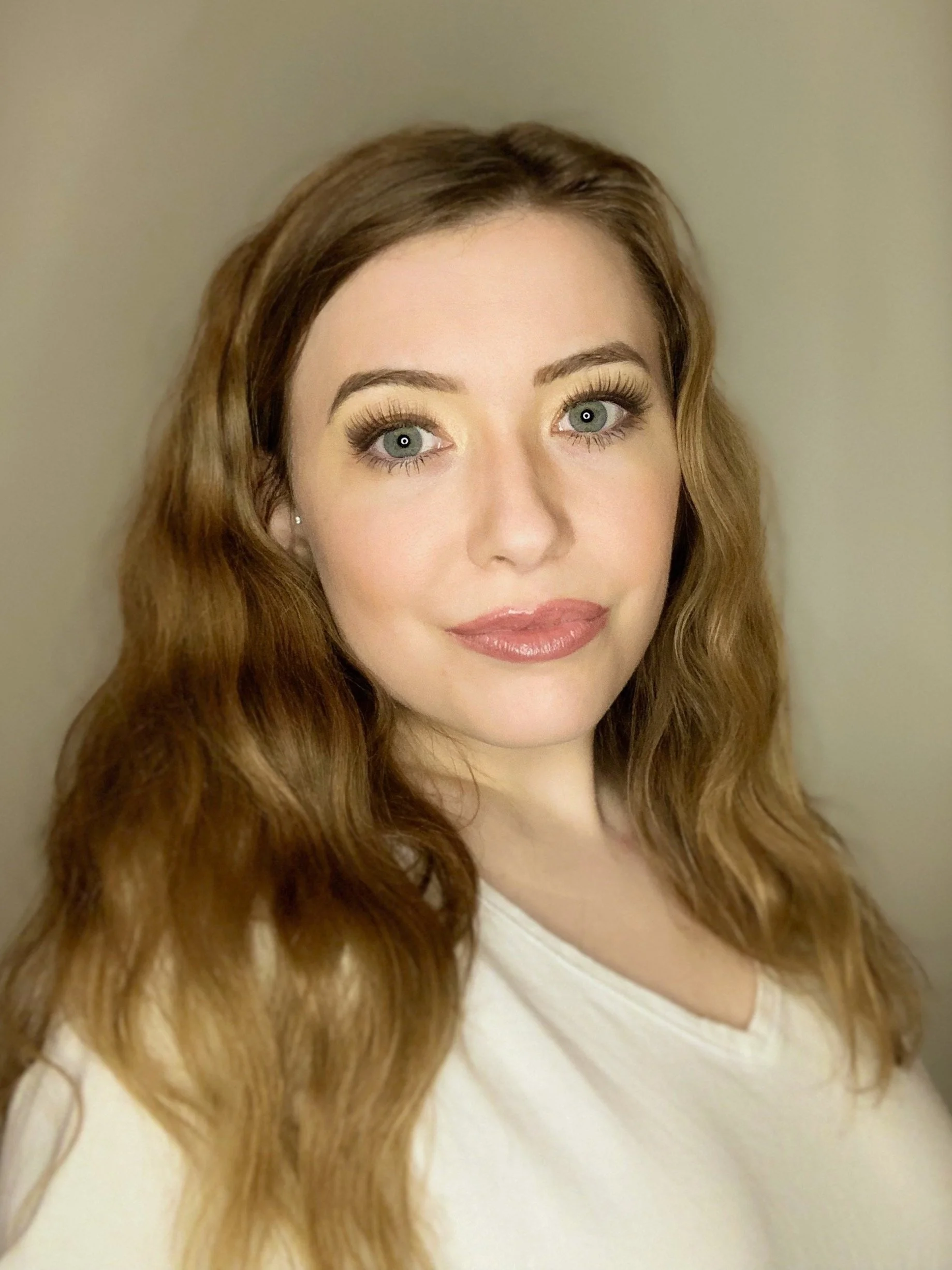 Woman with long wavy hair and a white shirt looking at the camera, neutral background.