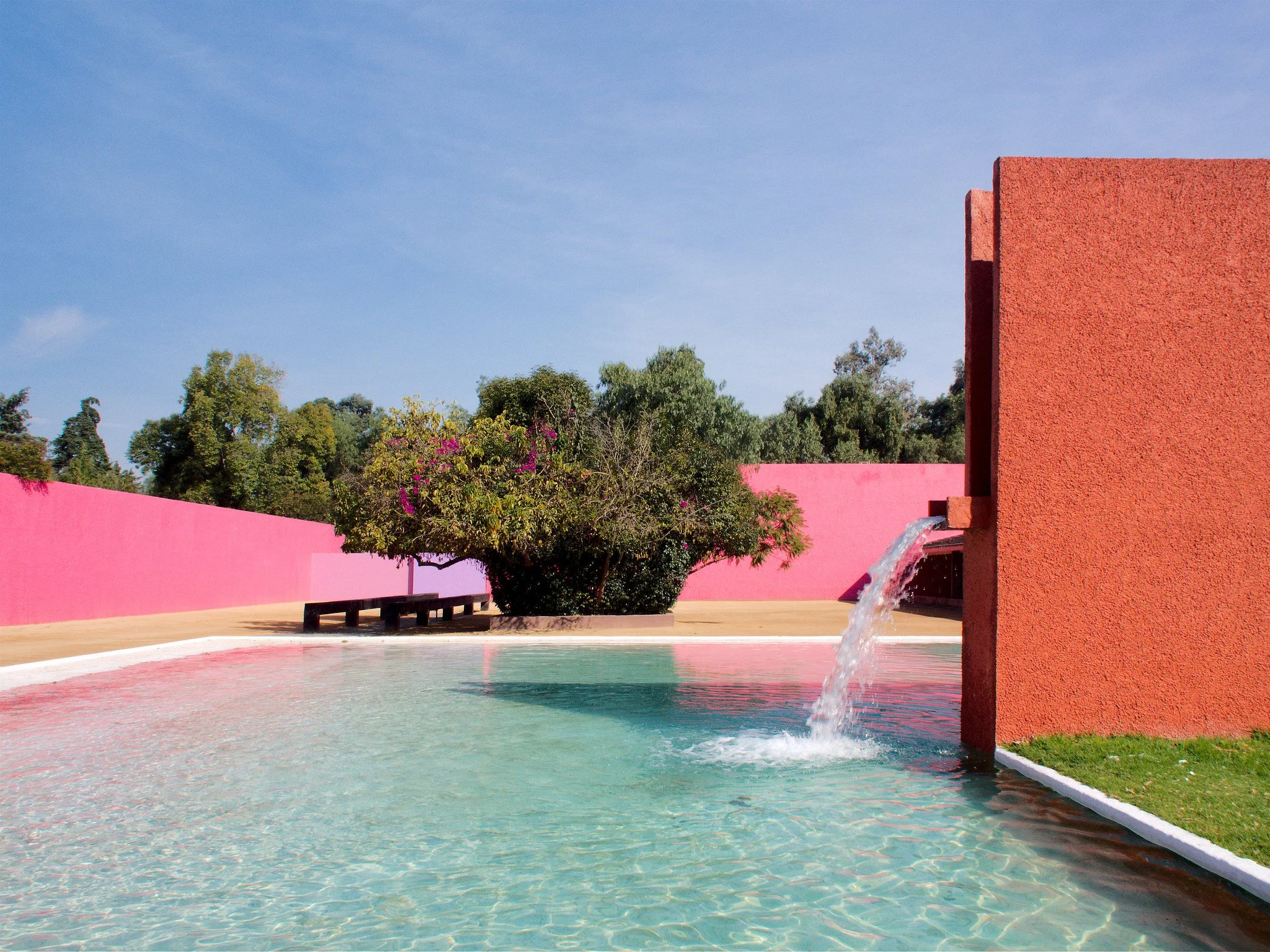 A spout juts from the side of an orange structure, feeding water into a pool. In the distance, block structures in pink line the pool. A backdrop of blue sky and green treetops frames the scene.