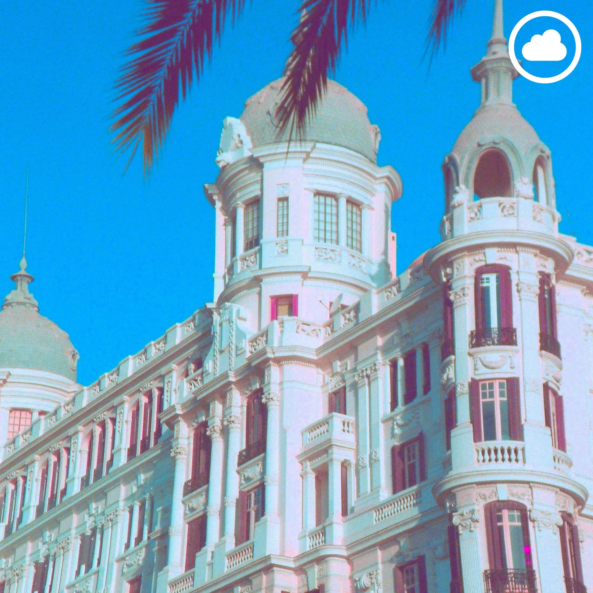 A white Victorian-style building with ornate architectural details and turrets, set against a bright blue sky, partly obscured by palm tree branches.