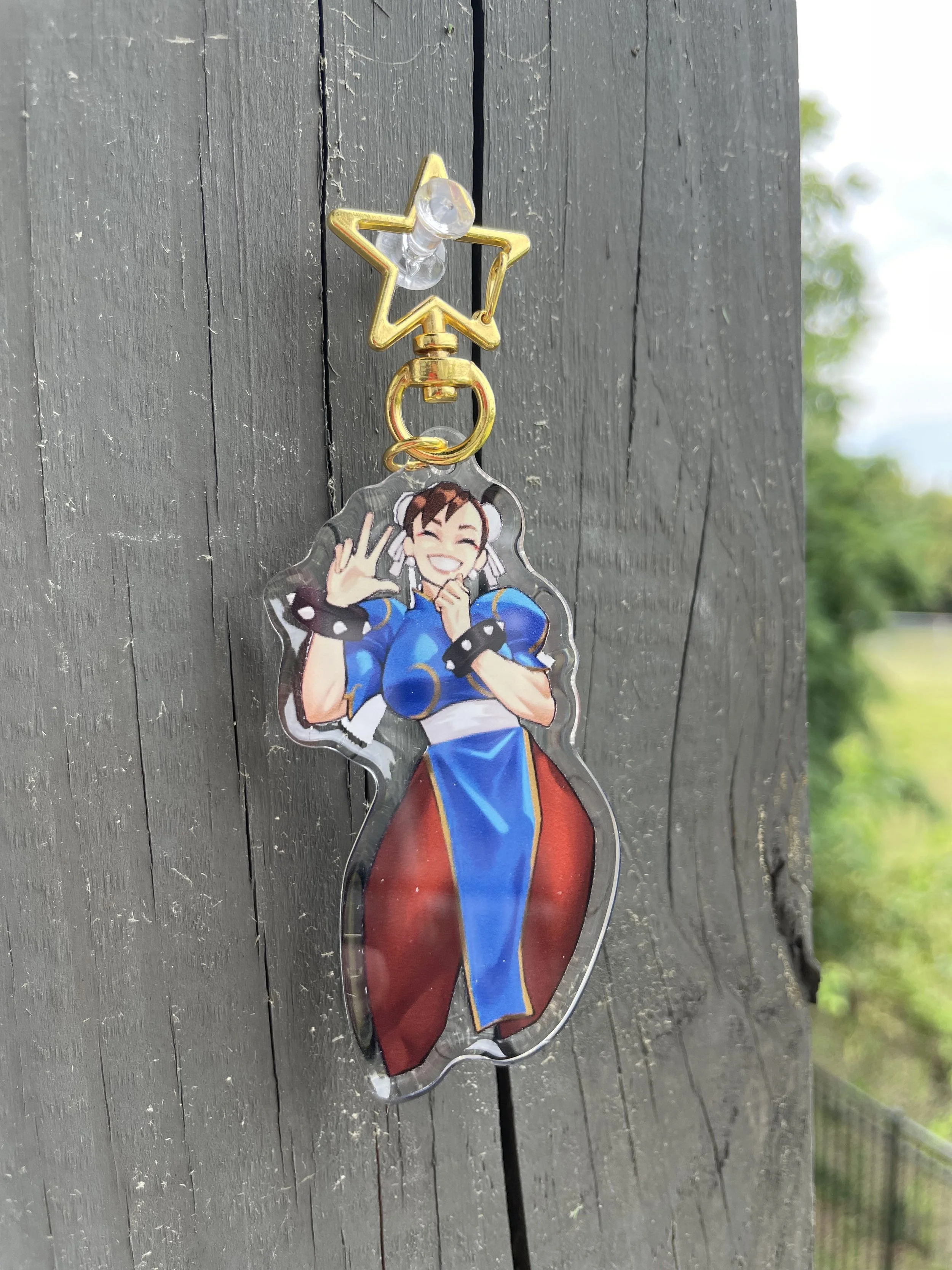 A keychain with a character art of a smiling girl wearing a blue and red outfit, making a peace sign with her right hand, attached to a gold star-shaped clip.