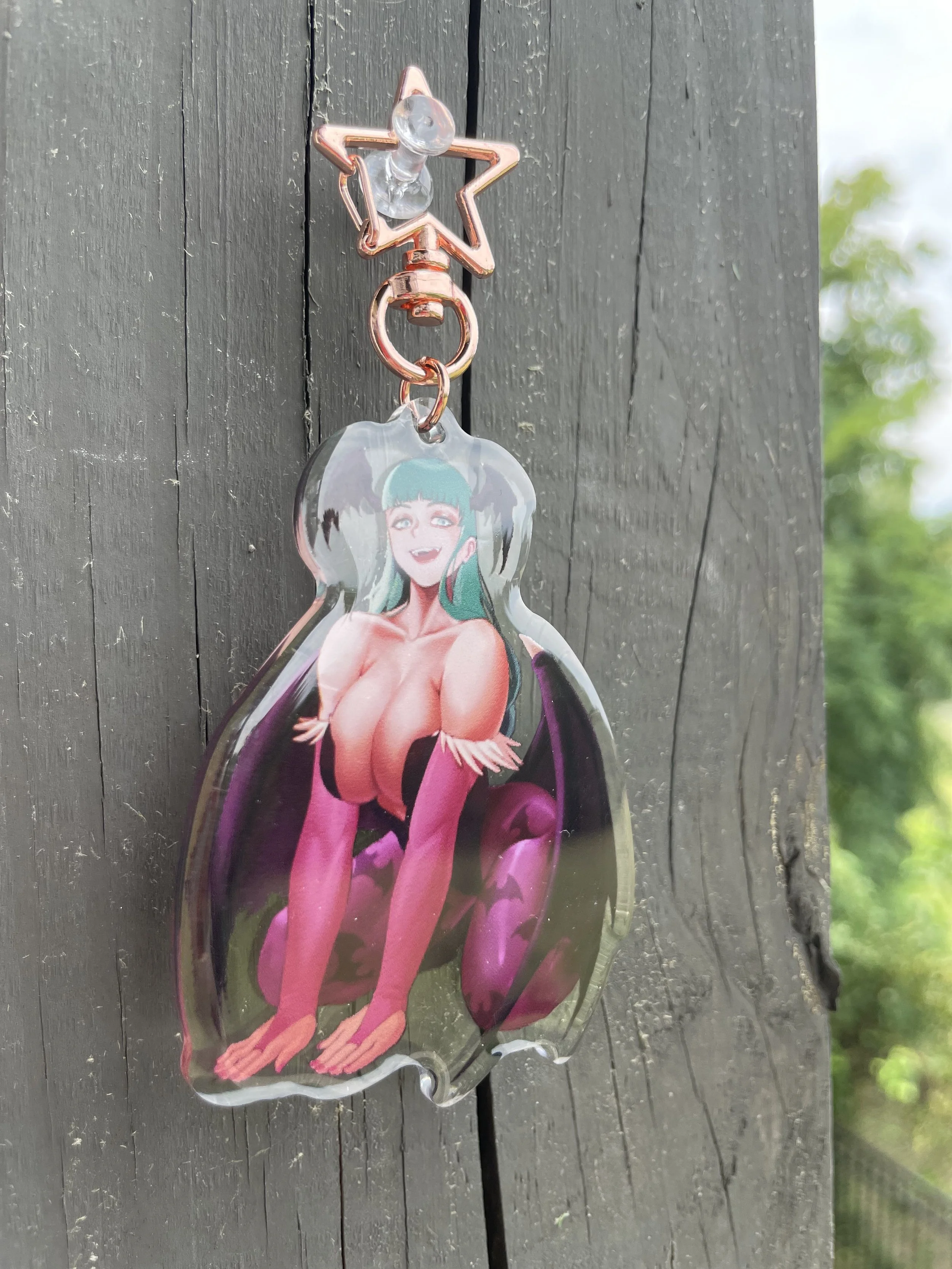 A keychain hanging on a dark wooden surface, featuring a colorful anime-style illustration of a smiling woman with green hair, wearing a revealing outfit with pink and purple stockings, and sitting on her knees with her hands on her knees.