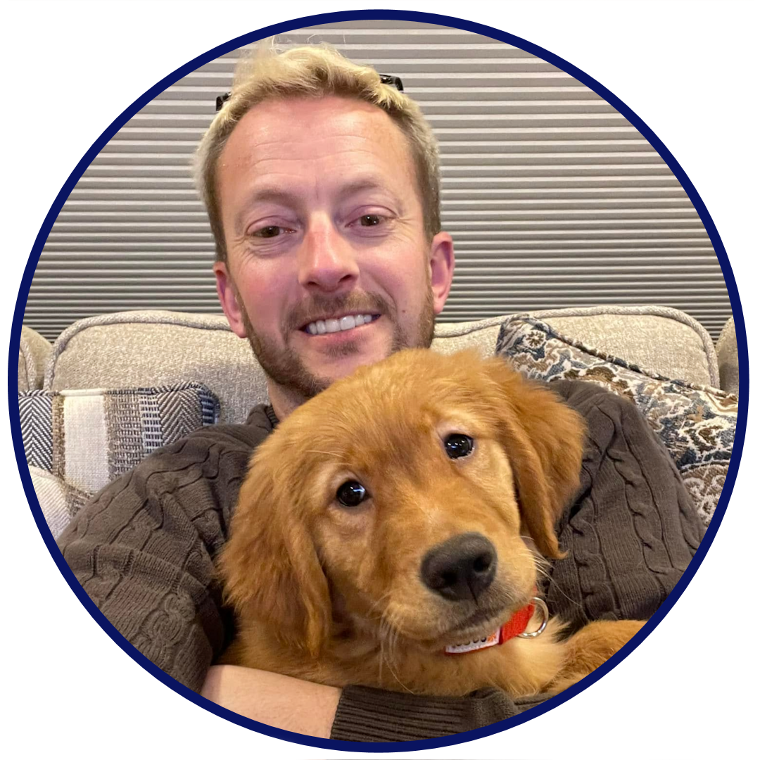 Town council candidate Christopher Peters sits on a couch facing the camera and holding his dog, Archie, who is also looking at the camera.