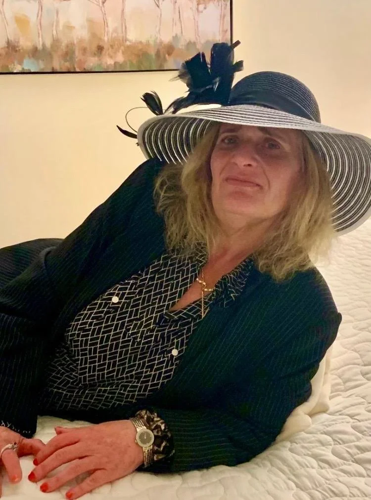 A woman lounging on a bed wearing a large striped black and white sun hat with decorative feathers, a dark pinstripe blazer, a patterned blouse, and jewelry including a watch, bracelet, and necklace.