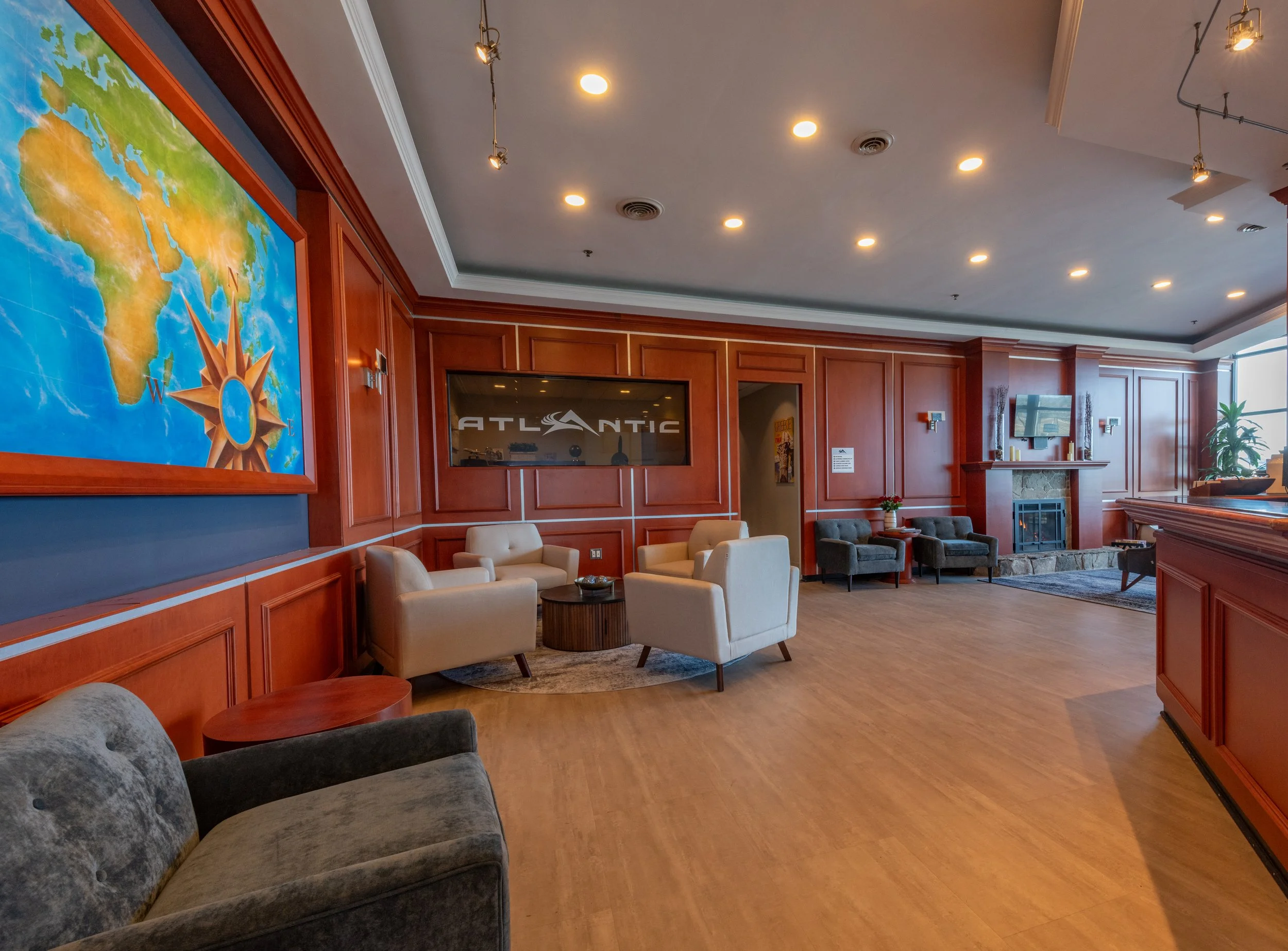 A spacious lobby area with wood-paneled walls, a marble fireplace, and comfortable seating including white and gray armchairs. A large world map is mounted on the wall to the left, and a flat-screen TV with the word "Atlantic" is visible. The ceiling has multiple recessed lights and track lighting, and a large window allows natural light into the room.