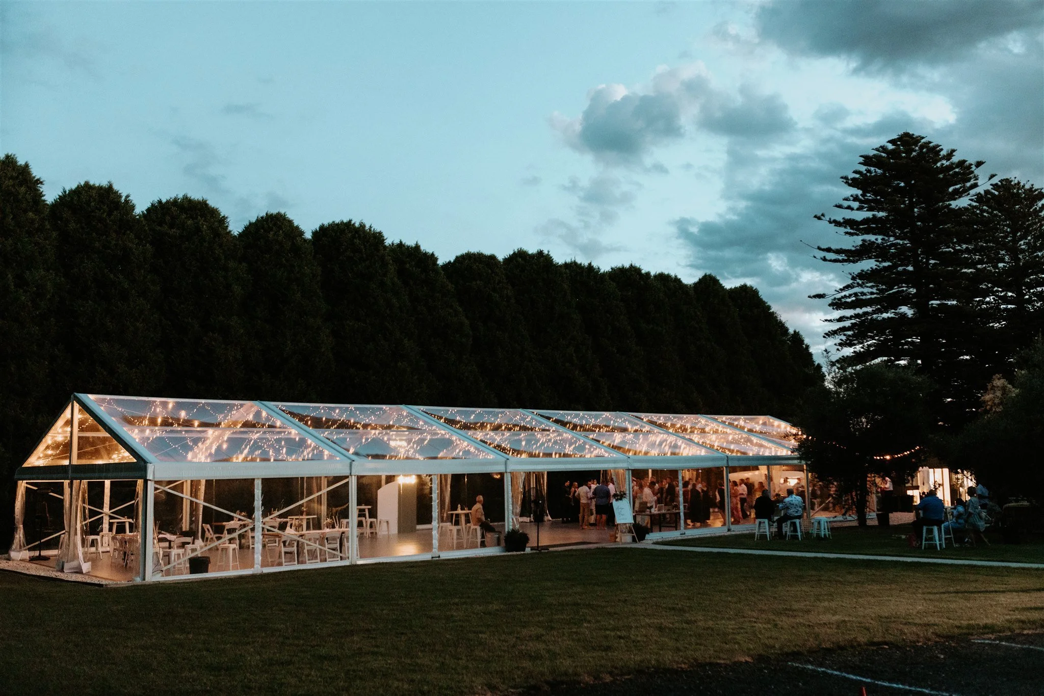 Outdoor event space with a clear tent decorated with string lights, set up on a grassy area during evening, with people gathering inside and outside.