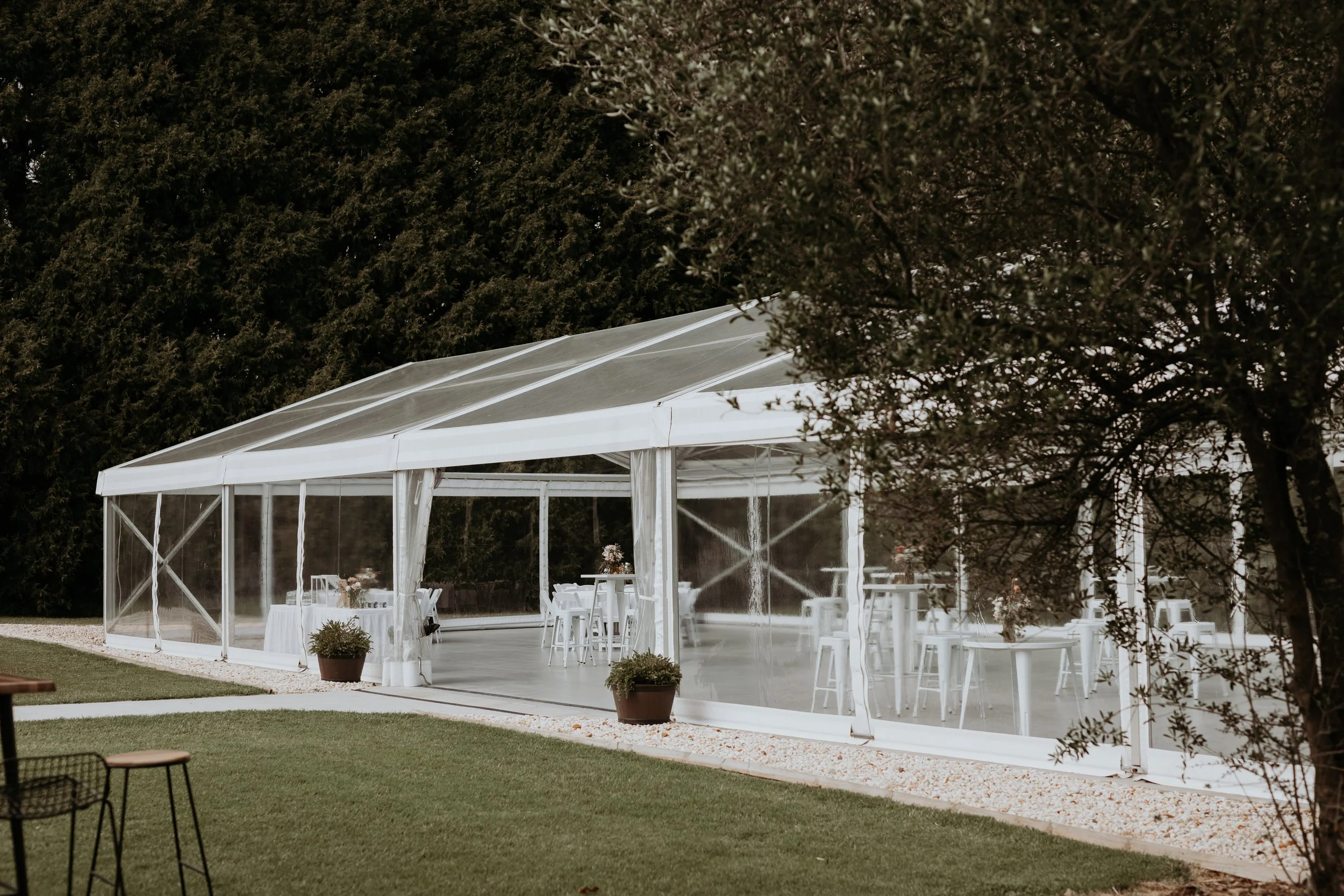White event tent set up outdoors on a grassy area, decorated with flowers and surrounded by trees.