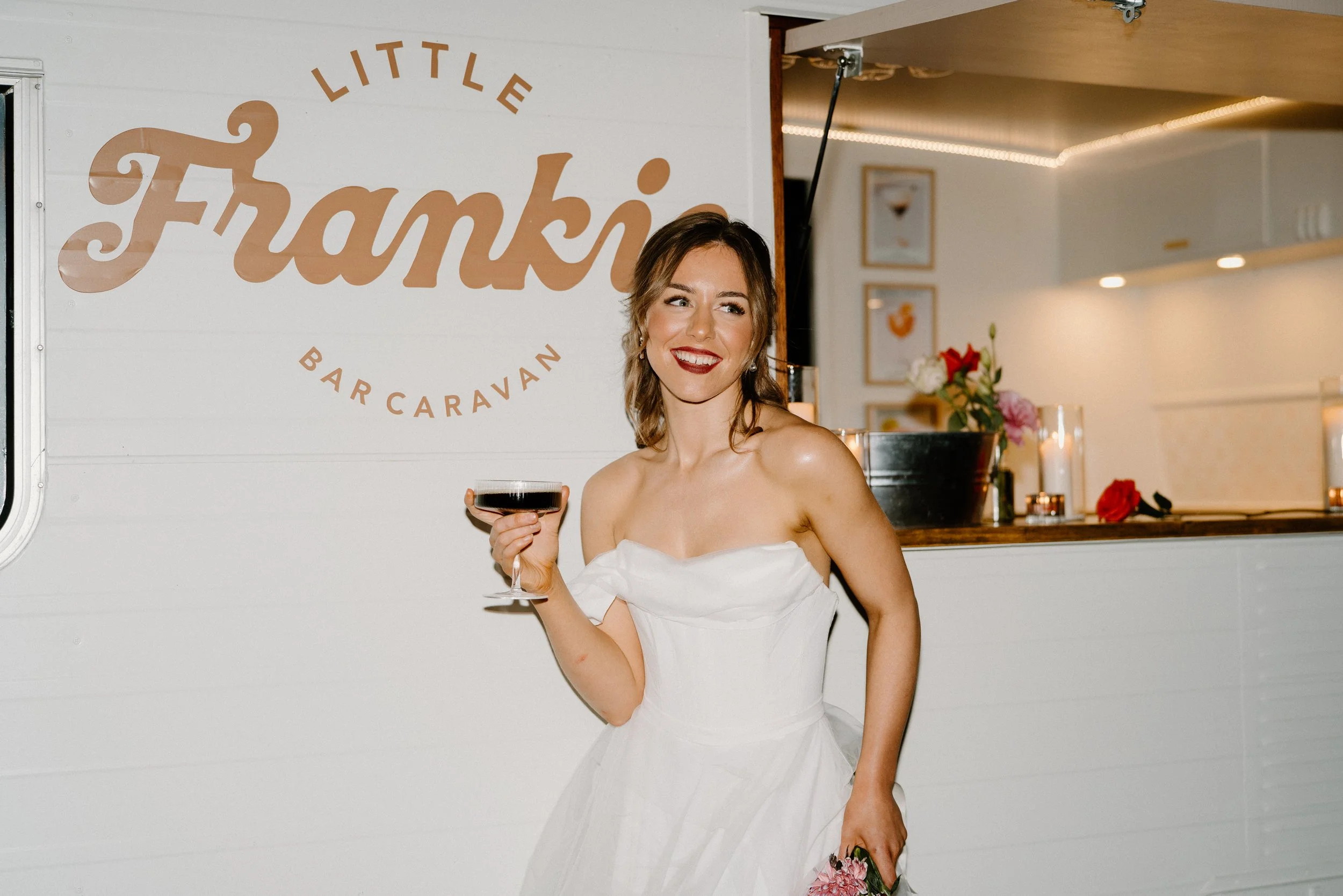 A woman in a white strapless dress holding a glass of red wine and smiling at an event at Little Frankie Bar Caravan.