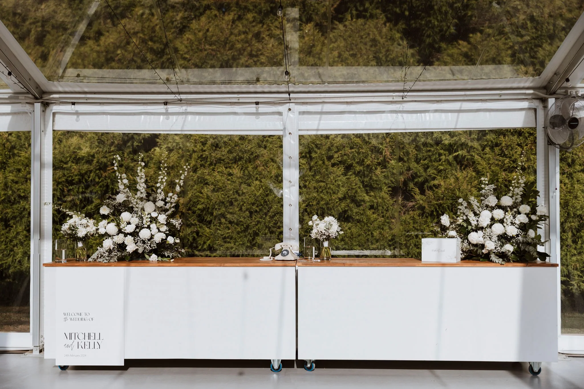 Wedding reception table decorated with large white floral arrangements, a vintage telephone, glass vases, and a white card on the right. The sign on the left reads 'Welcome to the wedding of Mitchel and Kelly, 24th February 2024.'