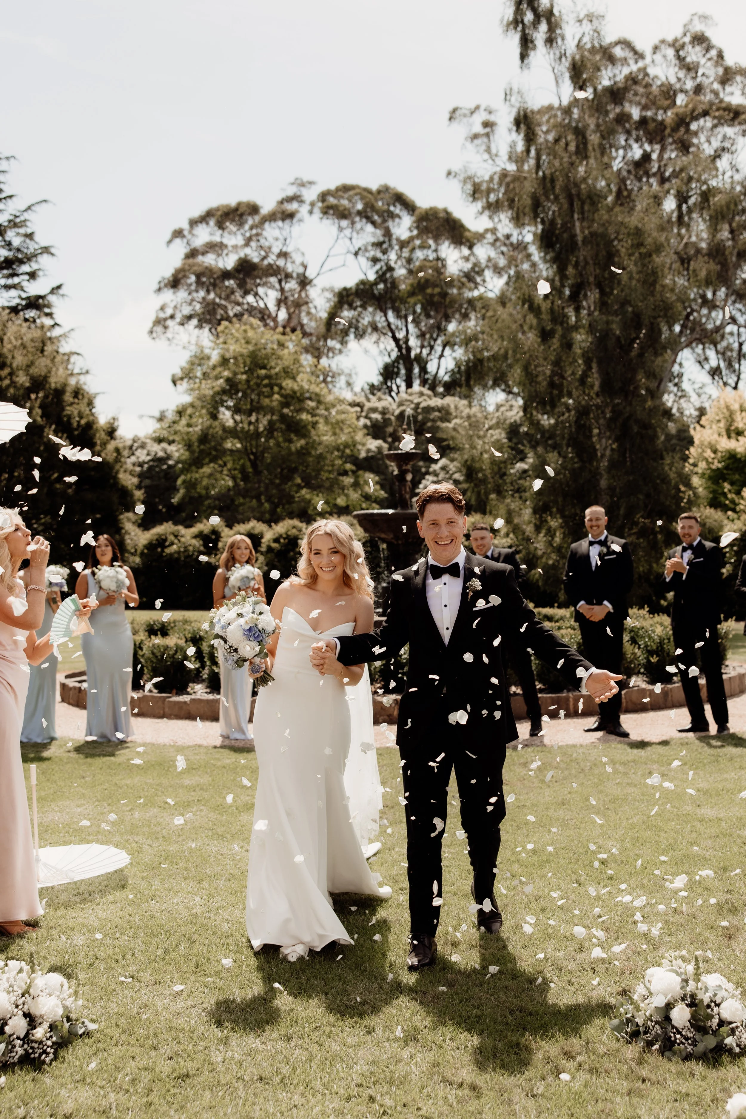 A newlywed couple smiling as they walk through falling flower petals in a garden, surrounded by friends and family celebrating their wedding.