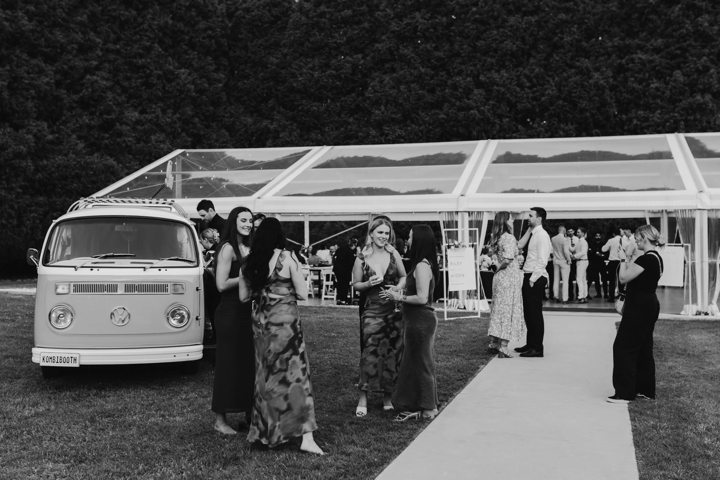 People gathered outside a transparent tent, with a vintage VW van parked nearby, at an outdoor event during daytime.