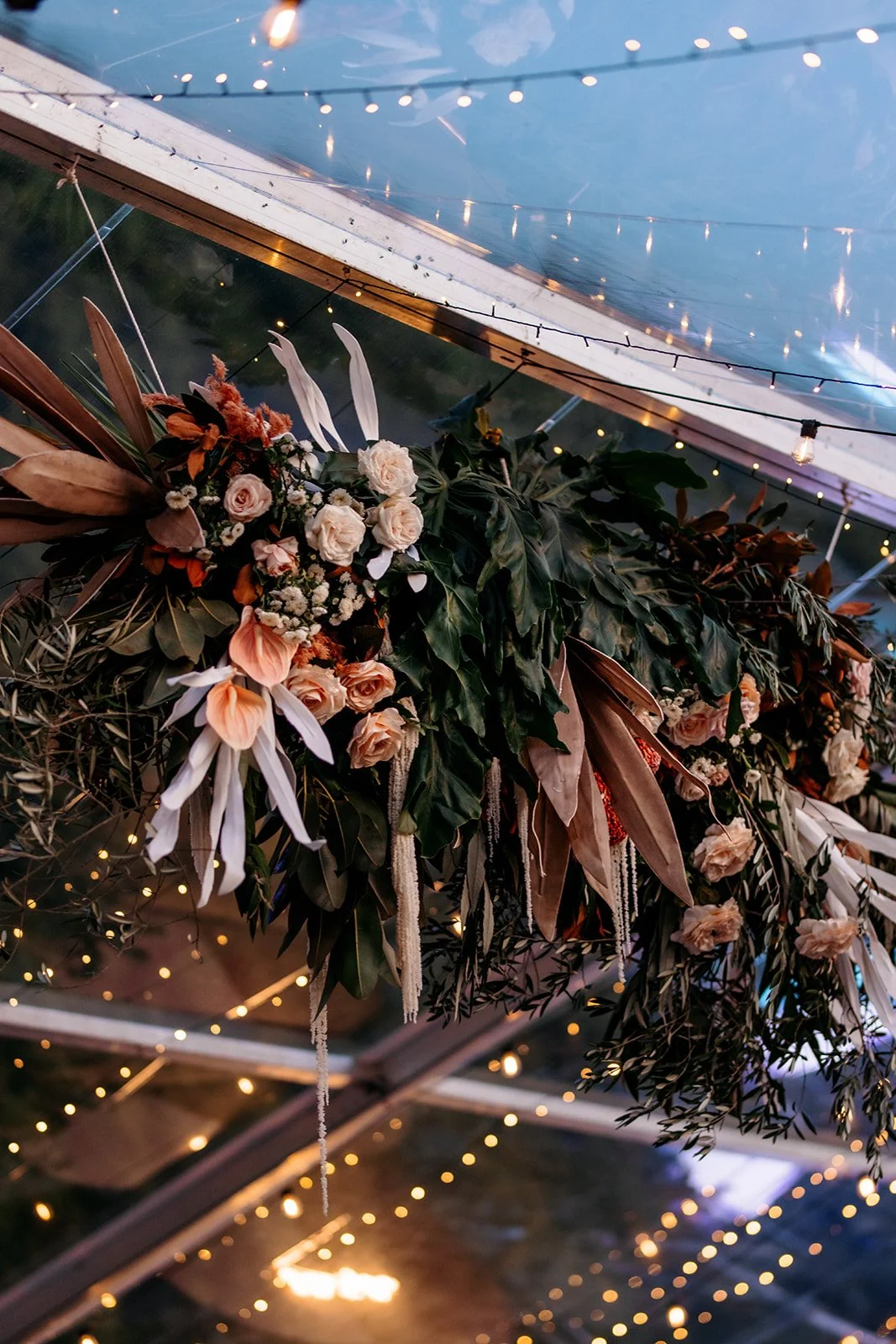 Floral and greenery arrangement hanging from ceiling with string lights and sky visible through transparent roof.