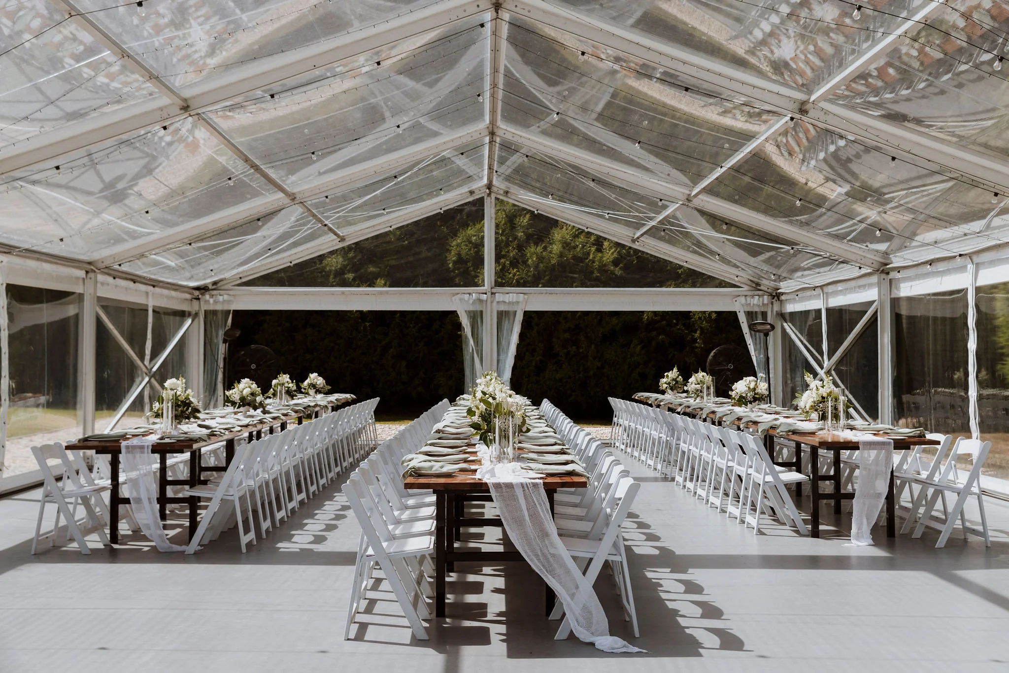 Long banquet table set up for an event inside a transparent tent, decorated with white flowers, linen table runners, and white chairs, with a backdrop of green trees.