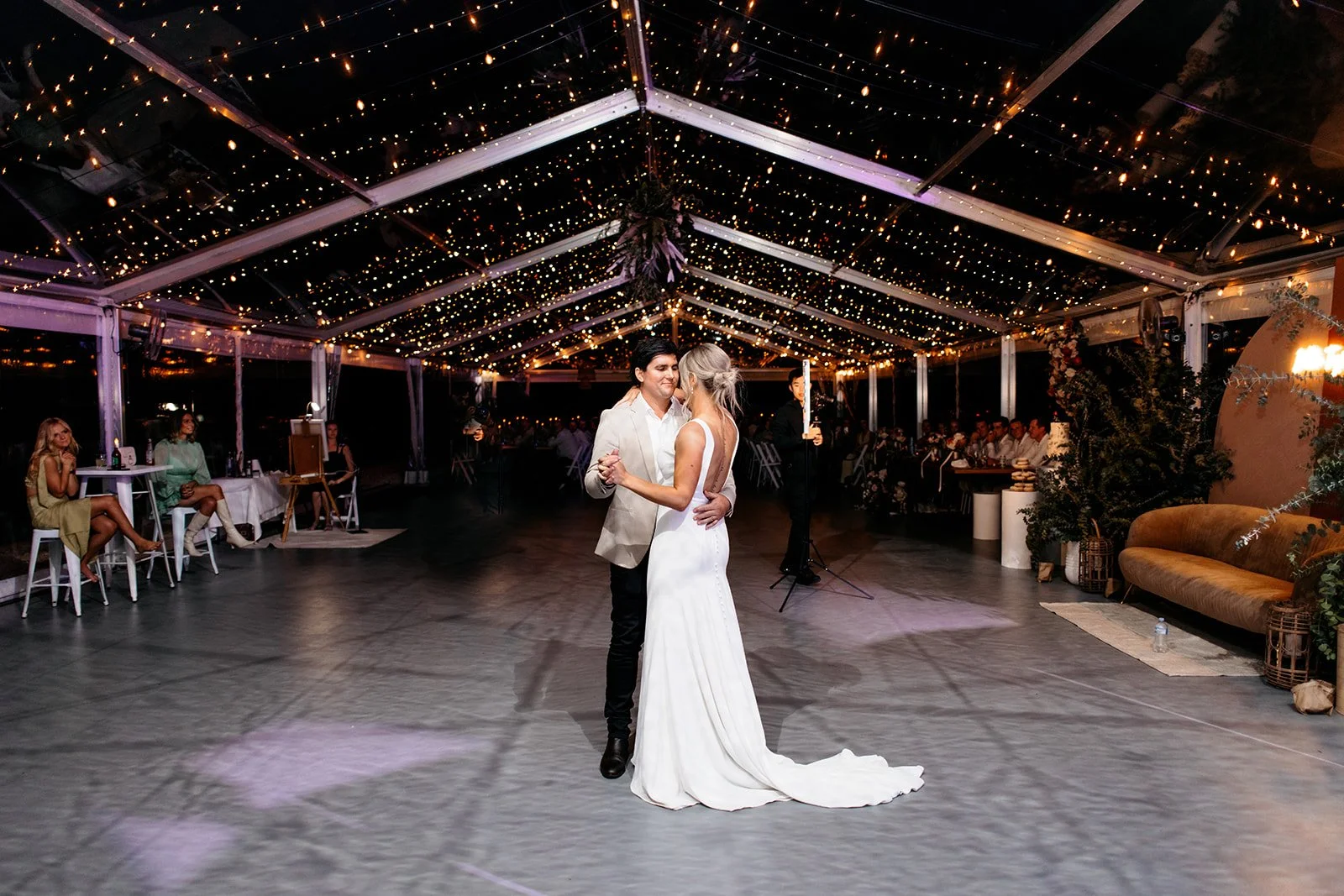 A couple dancing at a wedding reception under a canopy decorated with string lights, with guests seated at tables around the dance floor.