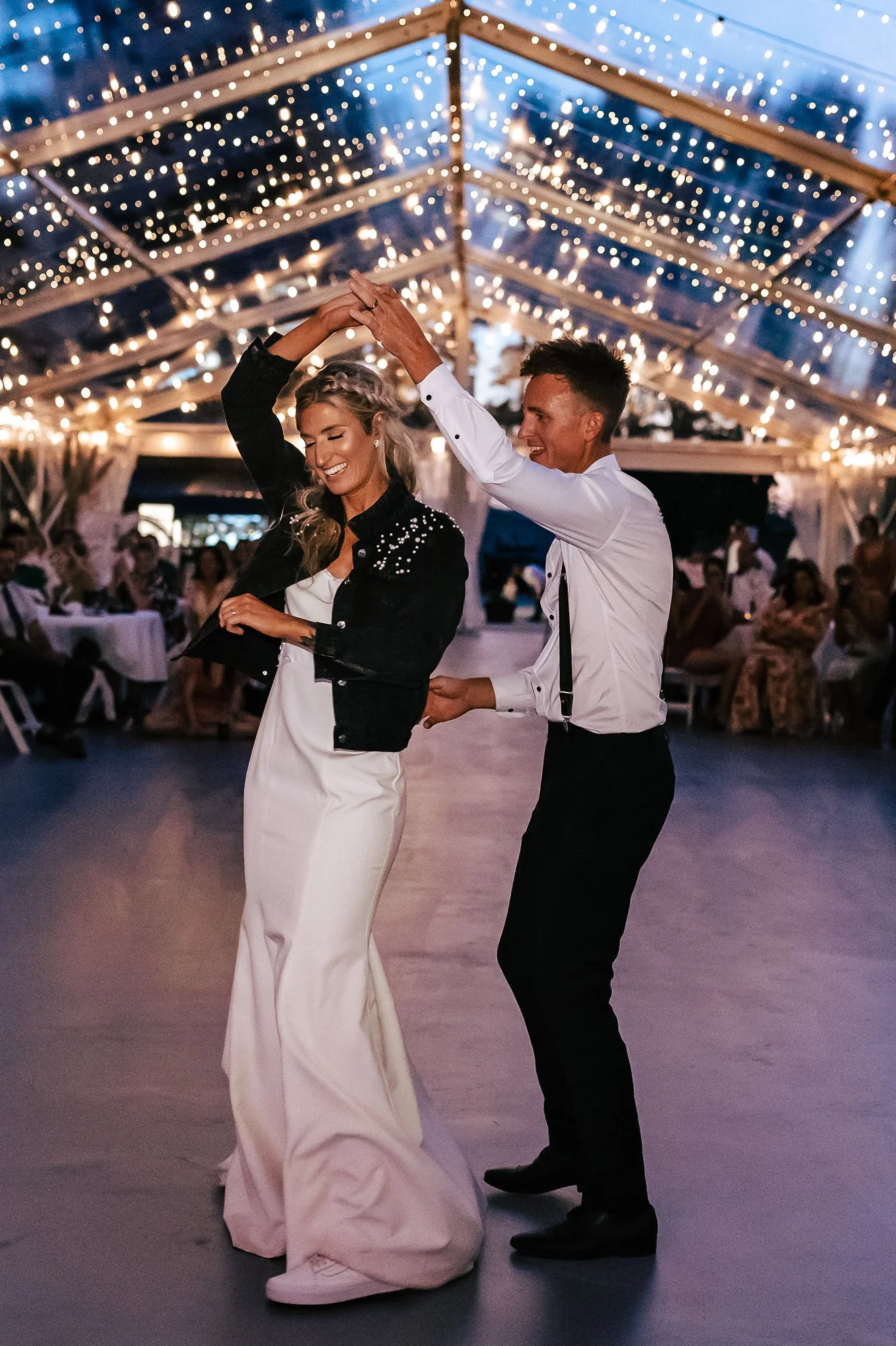 A couple dancing at a wedding reception under string lights inside a decorated tent, smiling and enjoying their dance.
