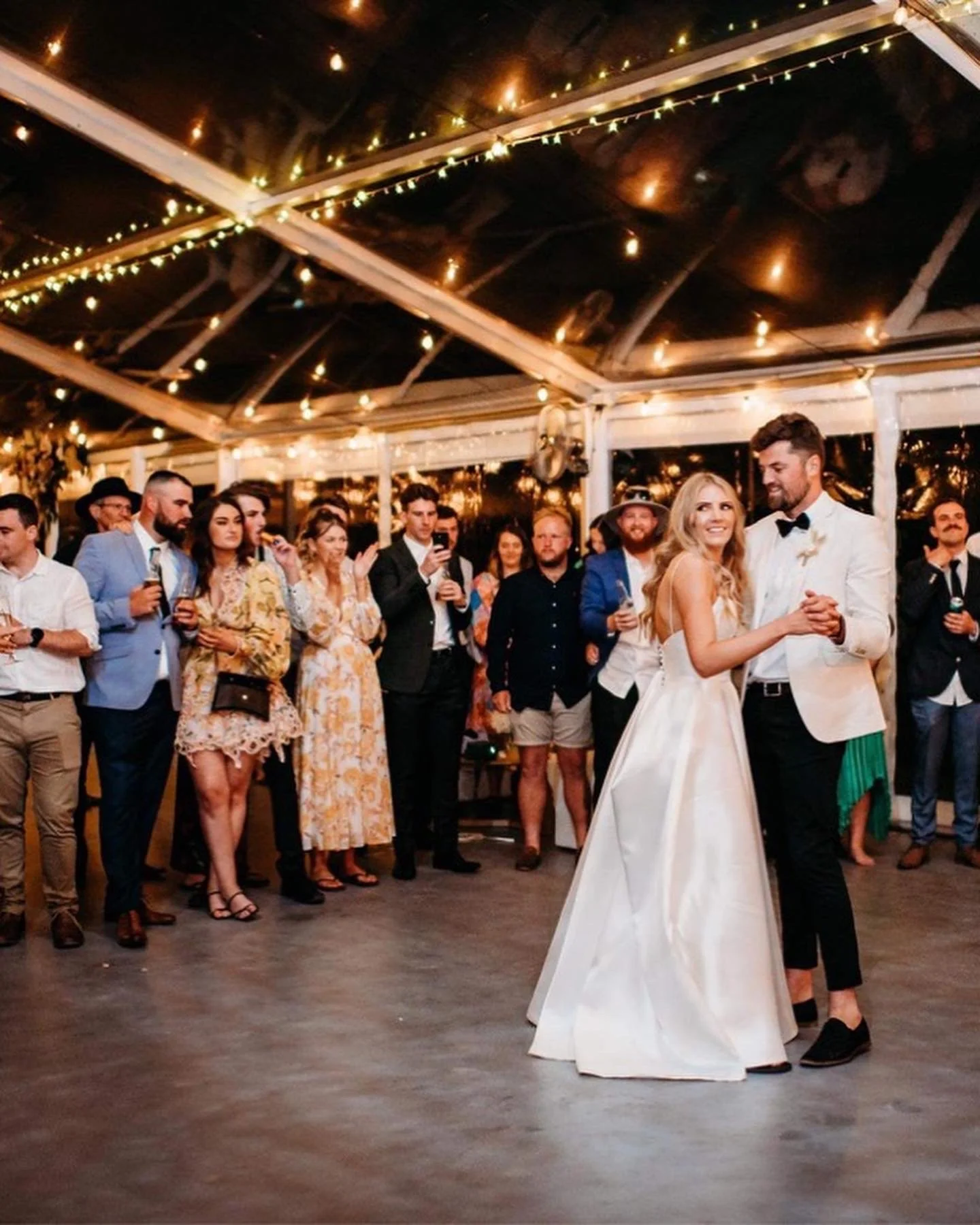 A bride and groom dance together at their wedding reception under string lights, with guests gathered around watching and celebrating.