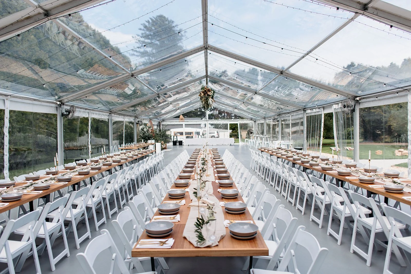 Long banquet tables set with plates, napkins, and silverware inside a transparent tent with string lights for an outdoor event.