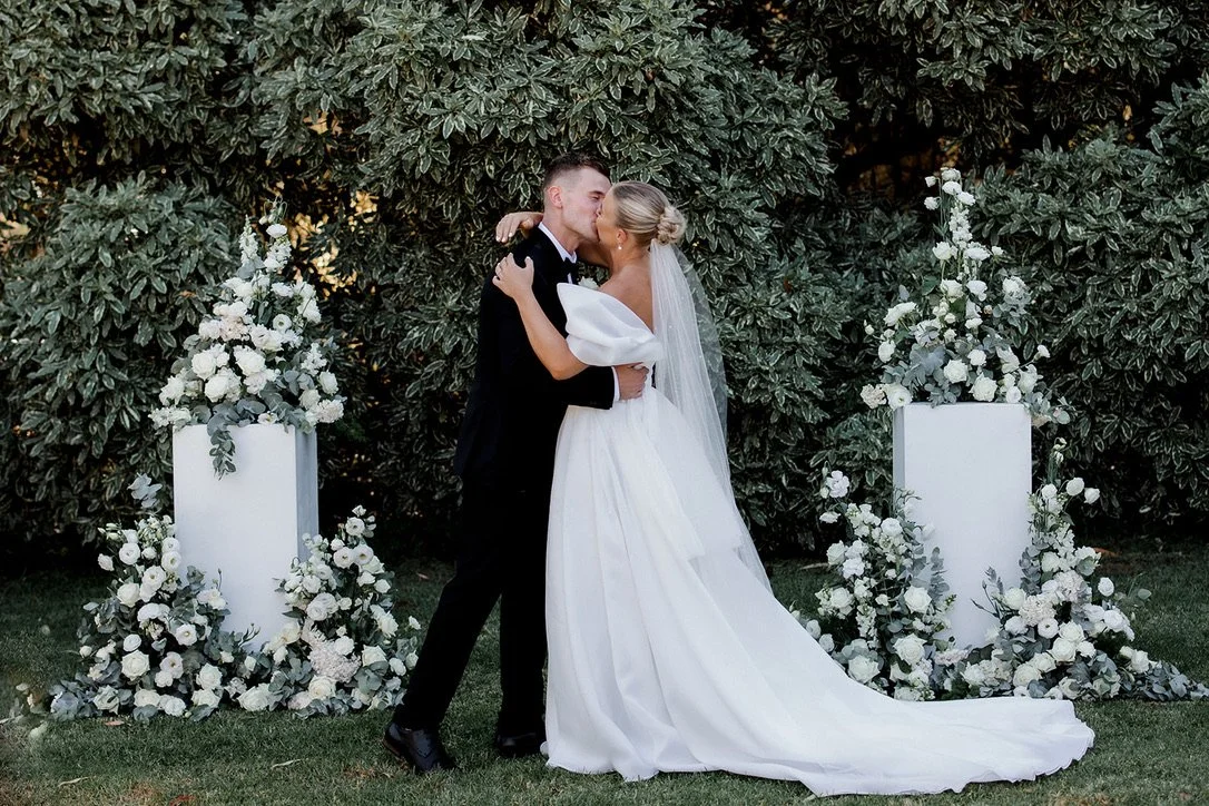 A bride and groom kissing outdoors with a backdrop of green bushes. The bride wears a white gown with puffed sleeves and a long veil. The groom wears a black suit. They are surrounded by white floral arrangements on white pedestals.