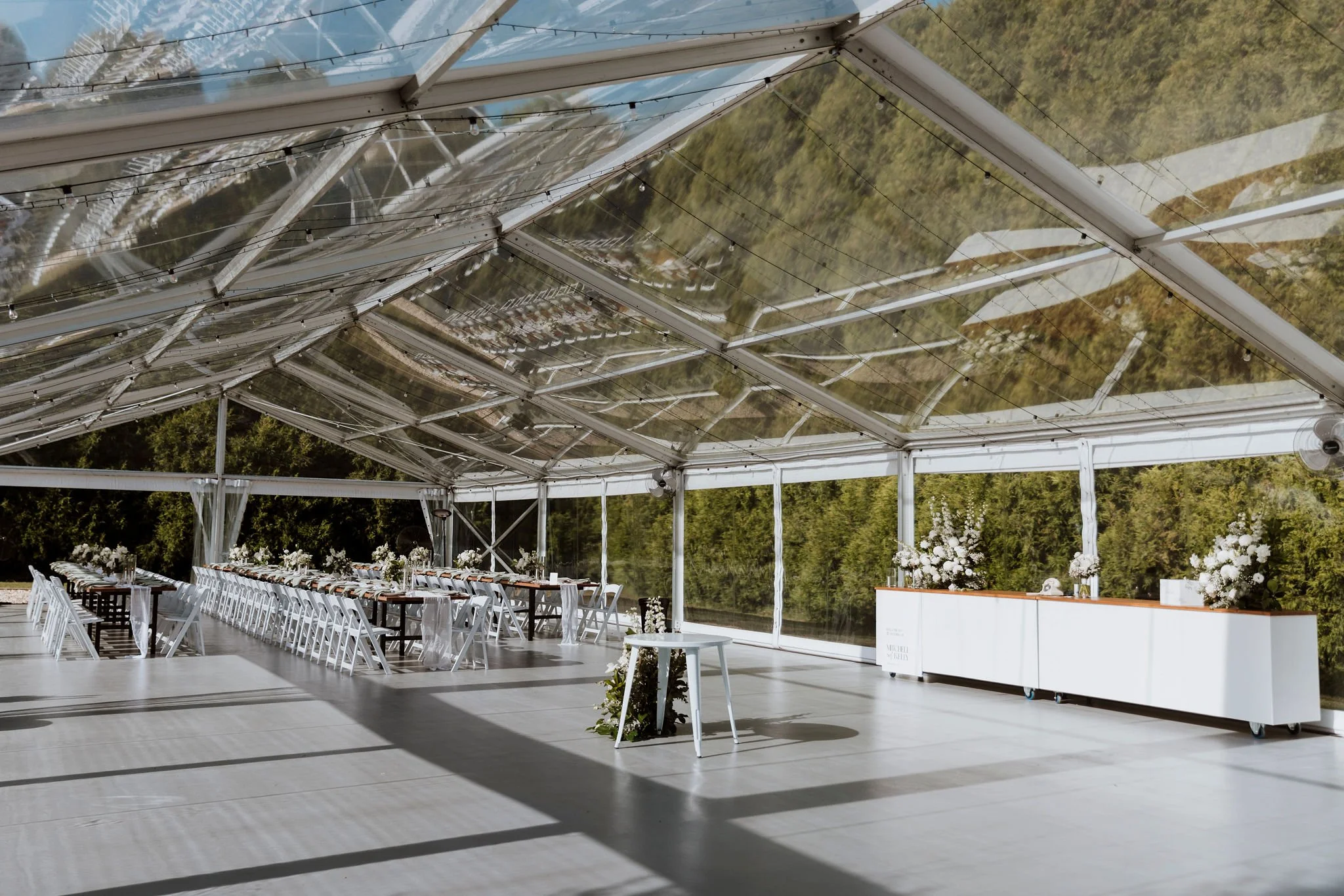 An empty event tent with white chairs and long tables decorated with floral arrangements, set up for a wedding or special event, with transparent walls and a view of greenery outside.