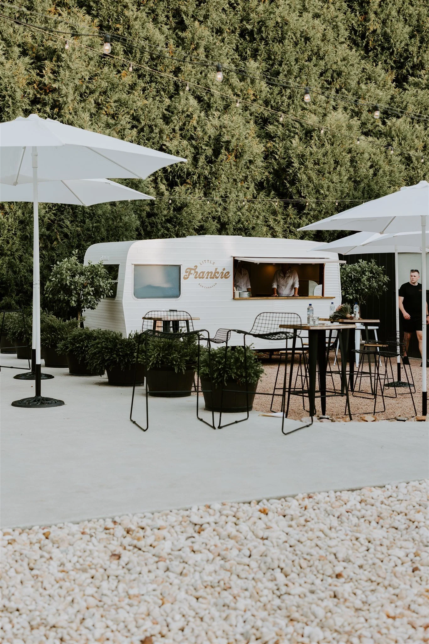 Outdoor seating area with black tables and chairs, white umbrellas, potted plants, and a vintage camper with a service window, set against a background of tall green trees.