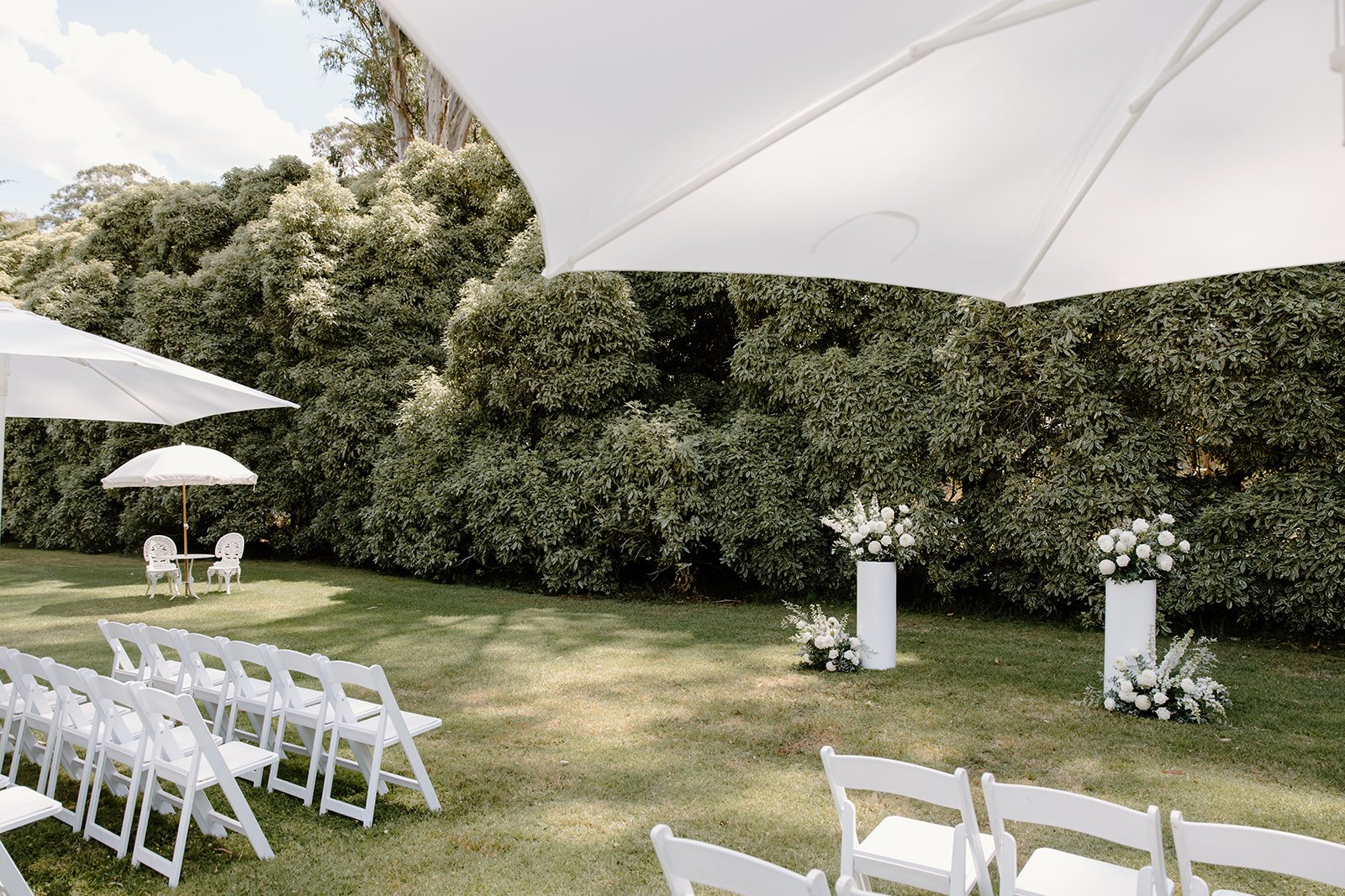 Outdoor wedding setup with white chairs, large white umbrellas, white floral arrangements on tall pedestals, and a grassy area surrounded by dense trees.