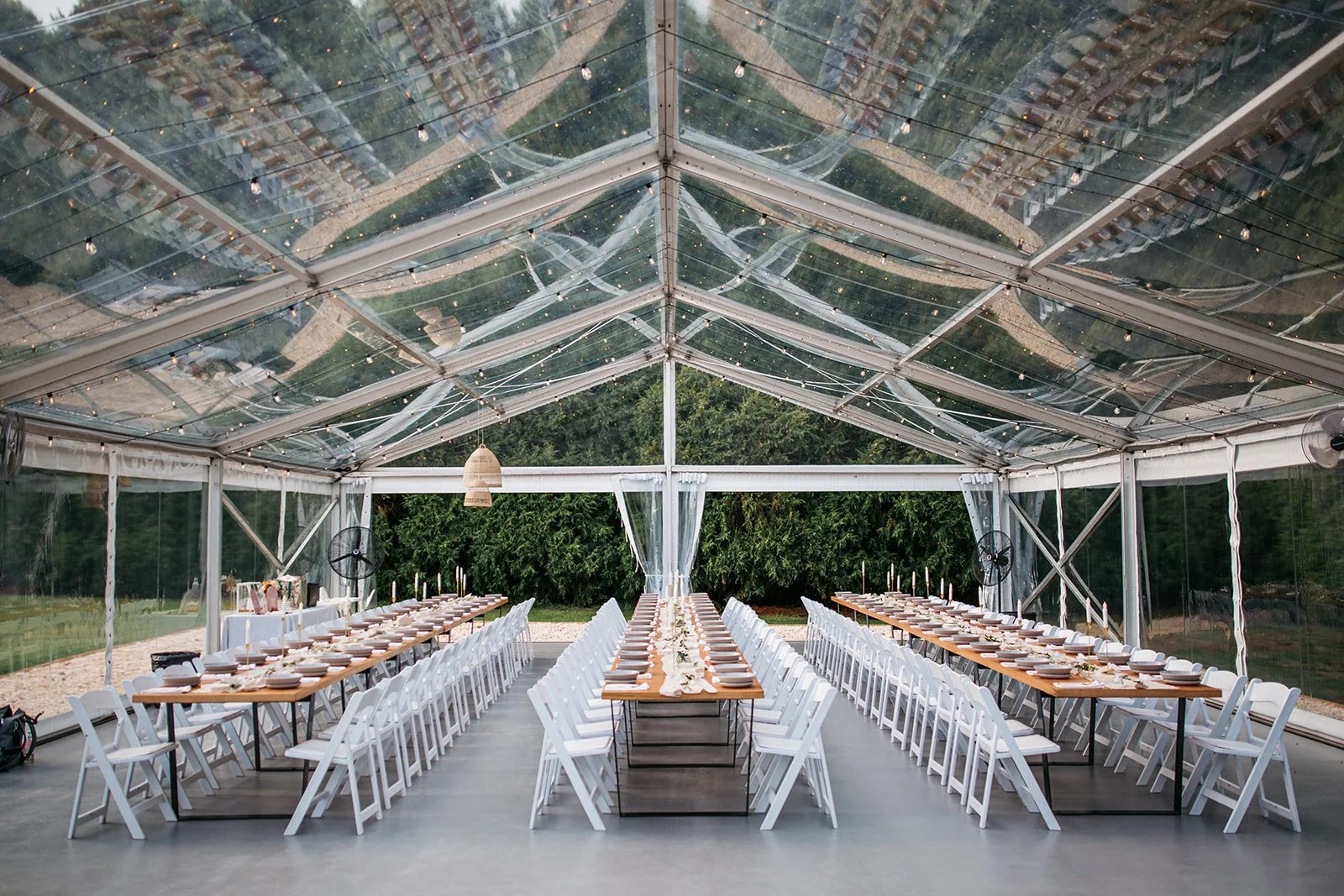 Elegant outdoor event space with long wooden tables set for dining, white chairs, inside a transparent tent with string lights and fans, overlooking a green landscape.