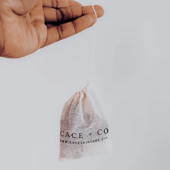 Hand holding a fabric drawstring bag with the text 'CACE + CO' and a website URL on it, against a plain light background.