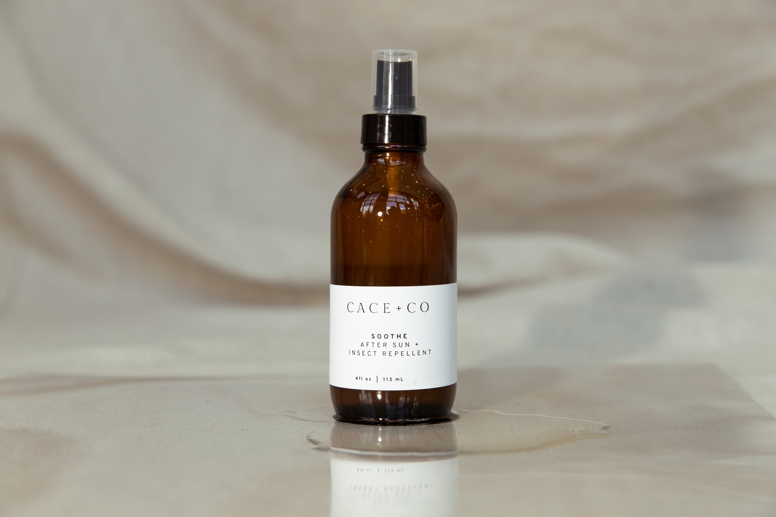 A brown glass spray bottle with a white label reading 'CACE + CO SOOTHE AFTER SUN + INSECT REPELLENT', sitting on a reflective surface with a light draped background.