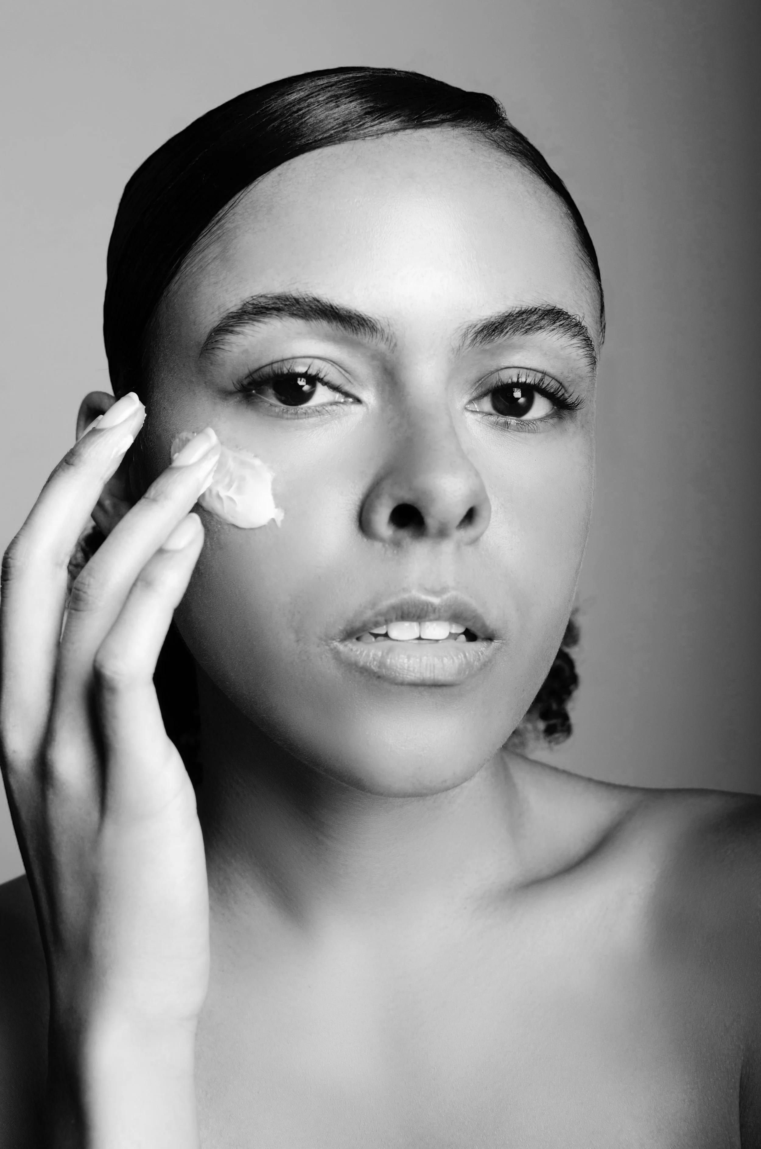 A black and white portrait of a woman with smooth skin applying cream to her face, looking directly at the camera.
