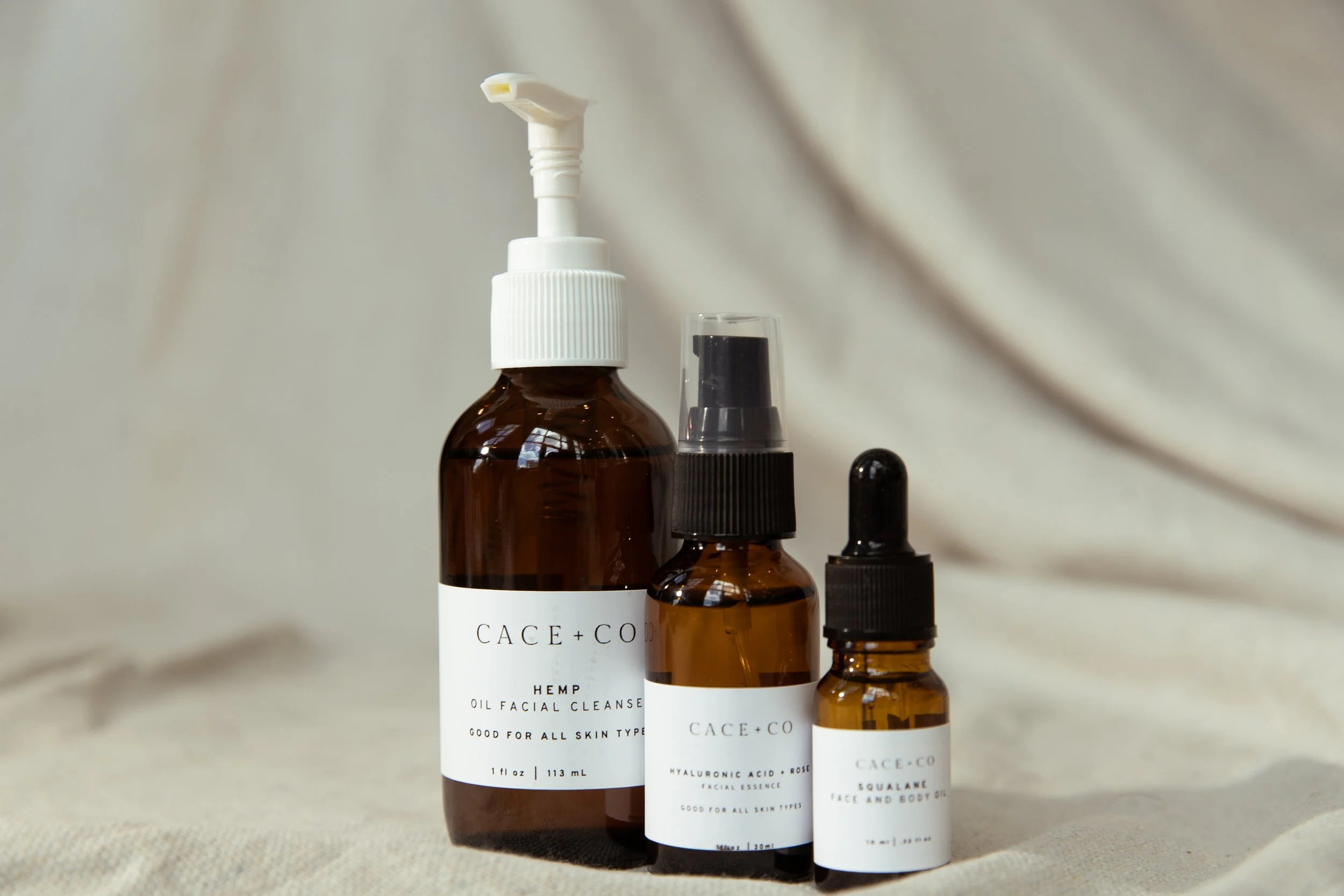 Three brown glass skincare bottles with white labels and black caps, labeled CACE+CO, containing facial cleansers and oils, set against a beige fabric background.