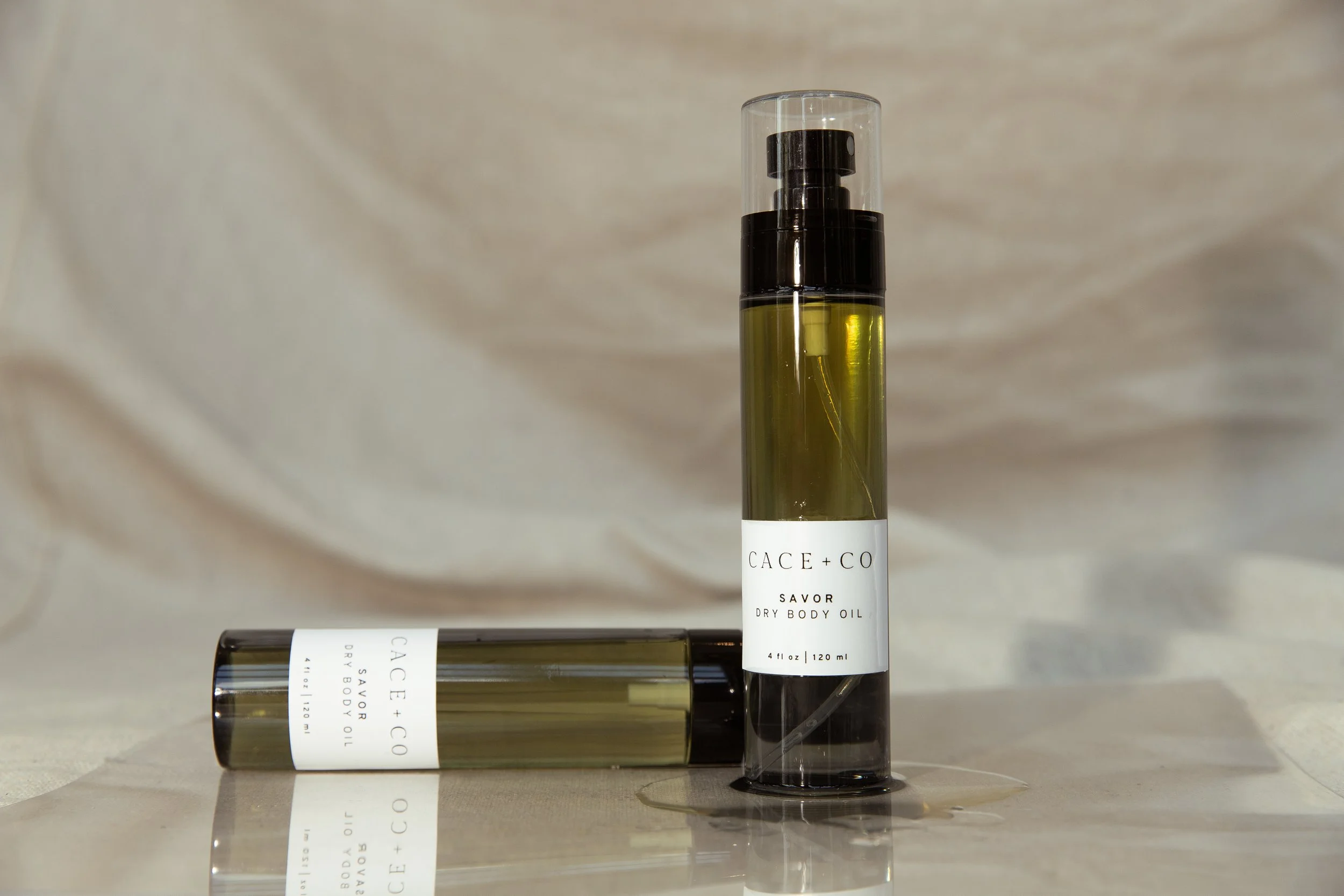 Two bottles of CACE+CO Savor Dry Body Oil, one standing upright and the other lying down, on a reflective surface with a draped fabric background.