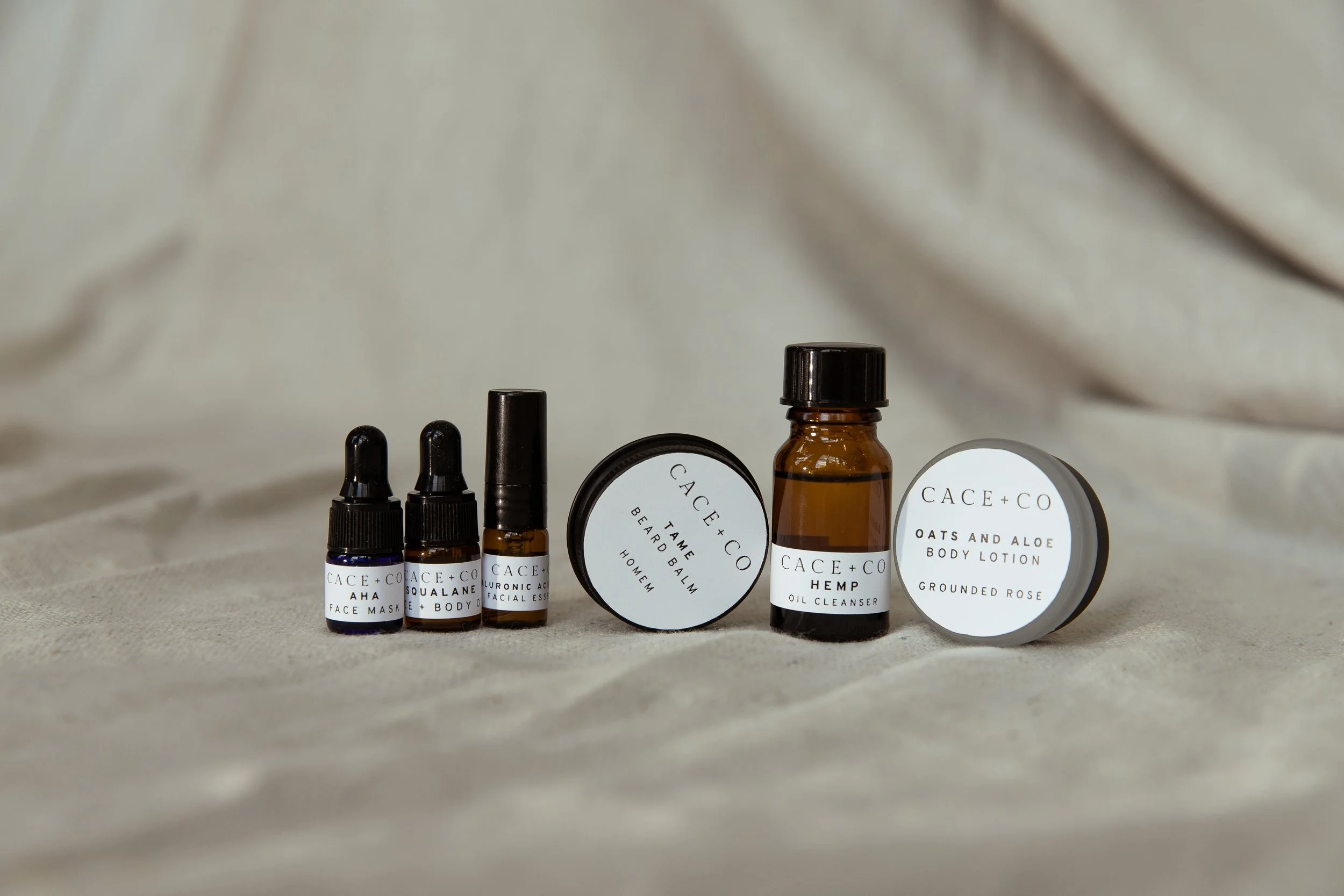 Collection of CACE + CO skincare products including face mask, Squalane + Body Oil, Tea Tree Beard Balm, Hemp Oil Cleanser, and Oats and Aloe Body Lotion displayed on fabric.