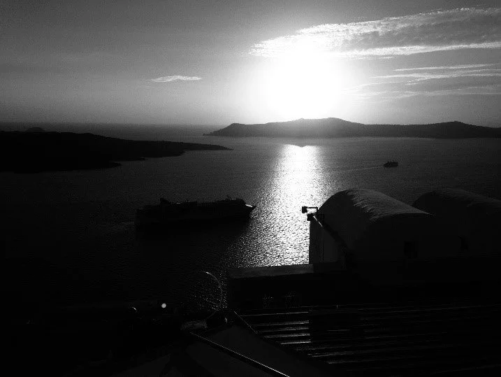 Black and white photo of a waterfront scene at sunset, with boats on the water and distant islands or mountains in the background.