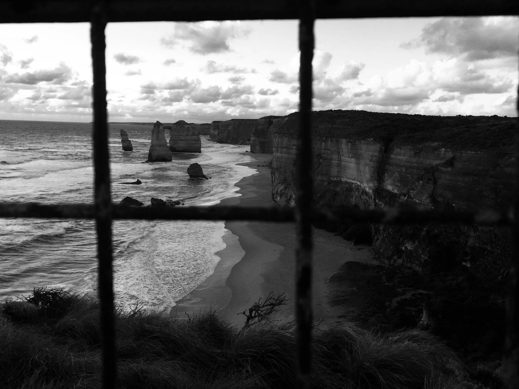 Black and white photograph of coastal cliffs with rock formations, ocean waves, and cloudy sky, viewed through a wire fence.