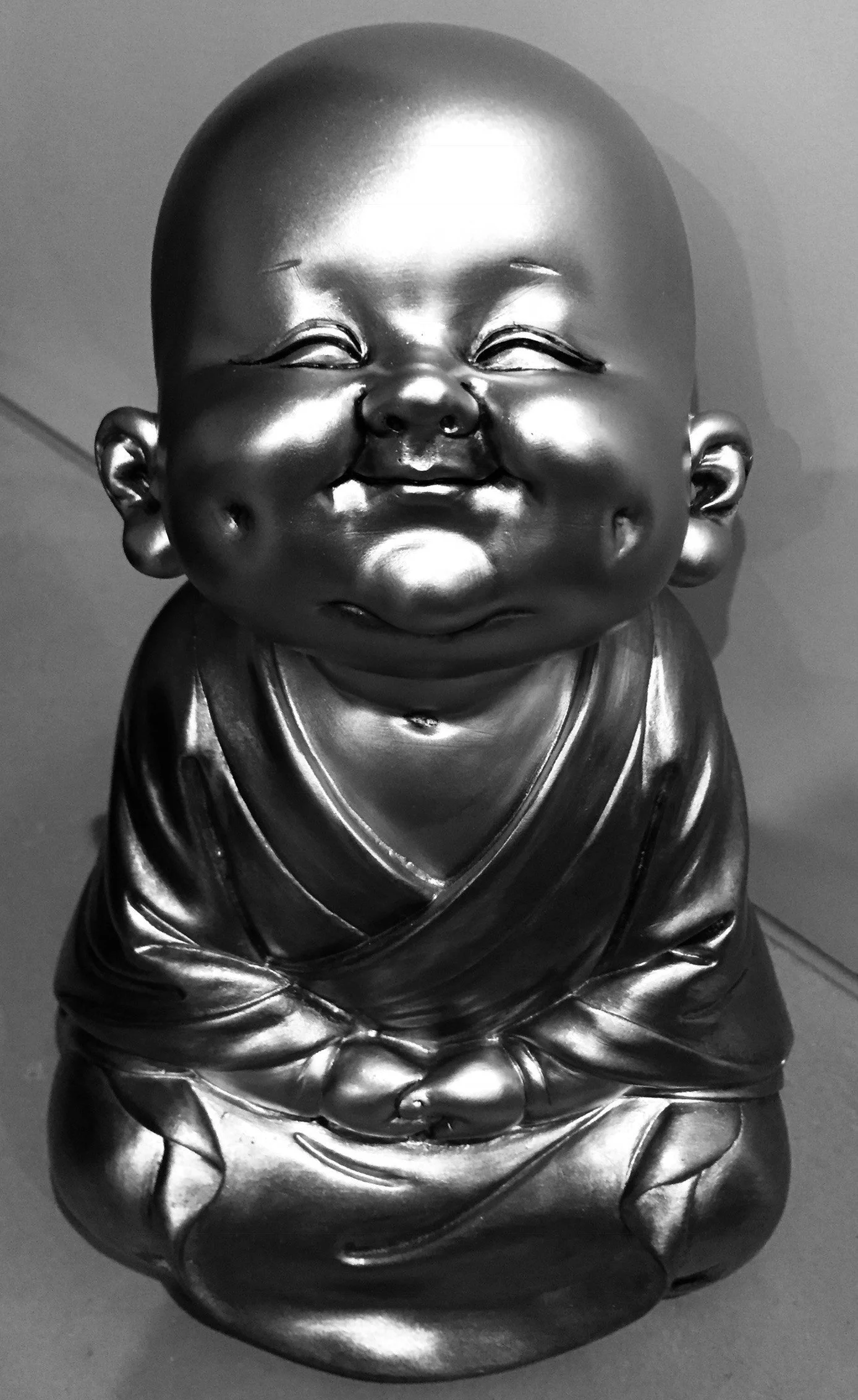 A black and white photo of a smiling statue of a young Buddhist monk in traditional robes, sitting cross-legged.