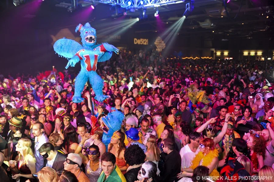 Large crowd of people at Carnaval Austin with colorful costumes and masks, and a large blue and red dragon puppet in the center.