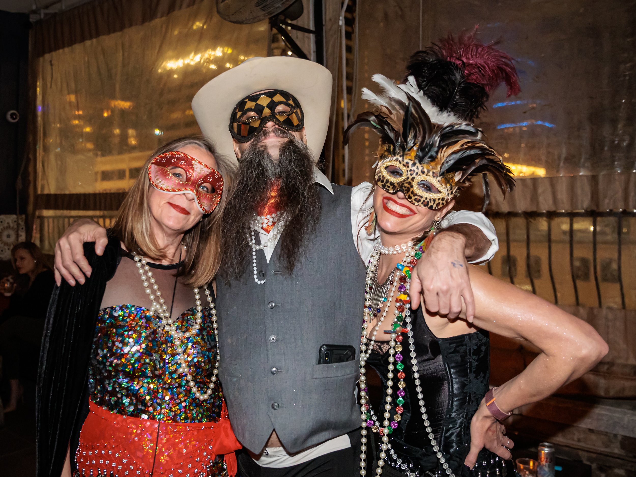 Three people at a party wearing colorful masks and costumes, smiling and hugging at Carnaval Brasileiro in Austin Texas, Returning February 7, 2026.